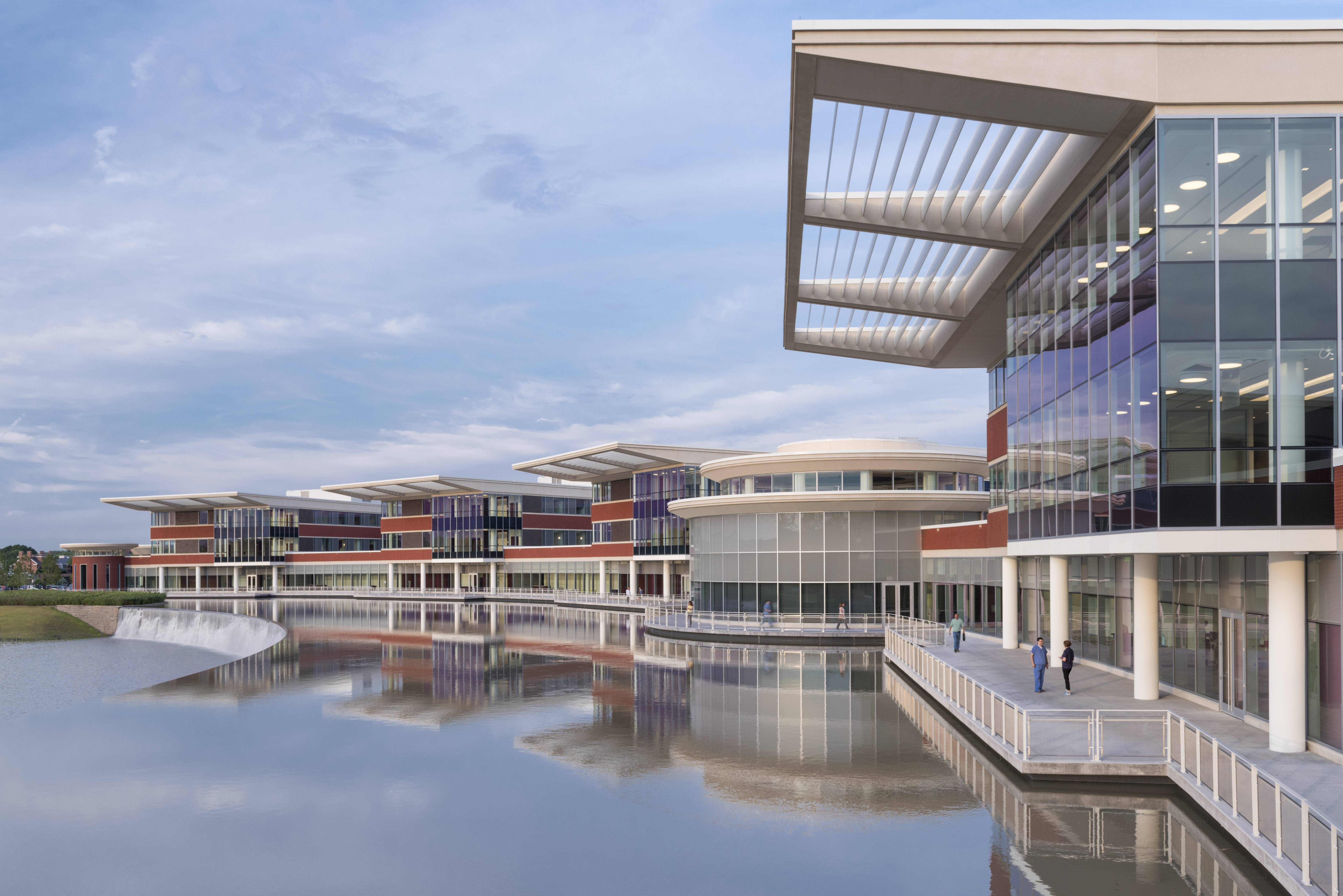 Modern multi-story hospital building with glass facade and canopy overhangs beside a reflective pond.