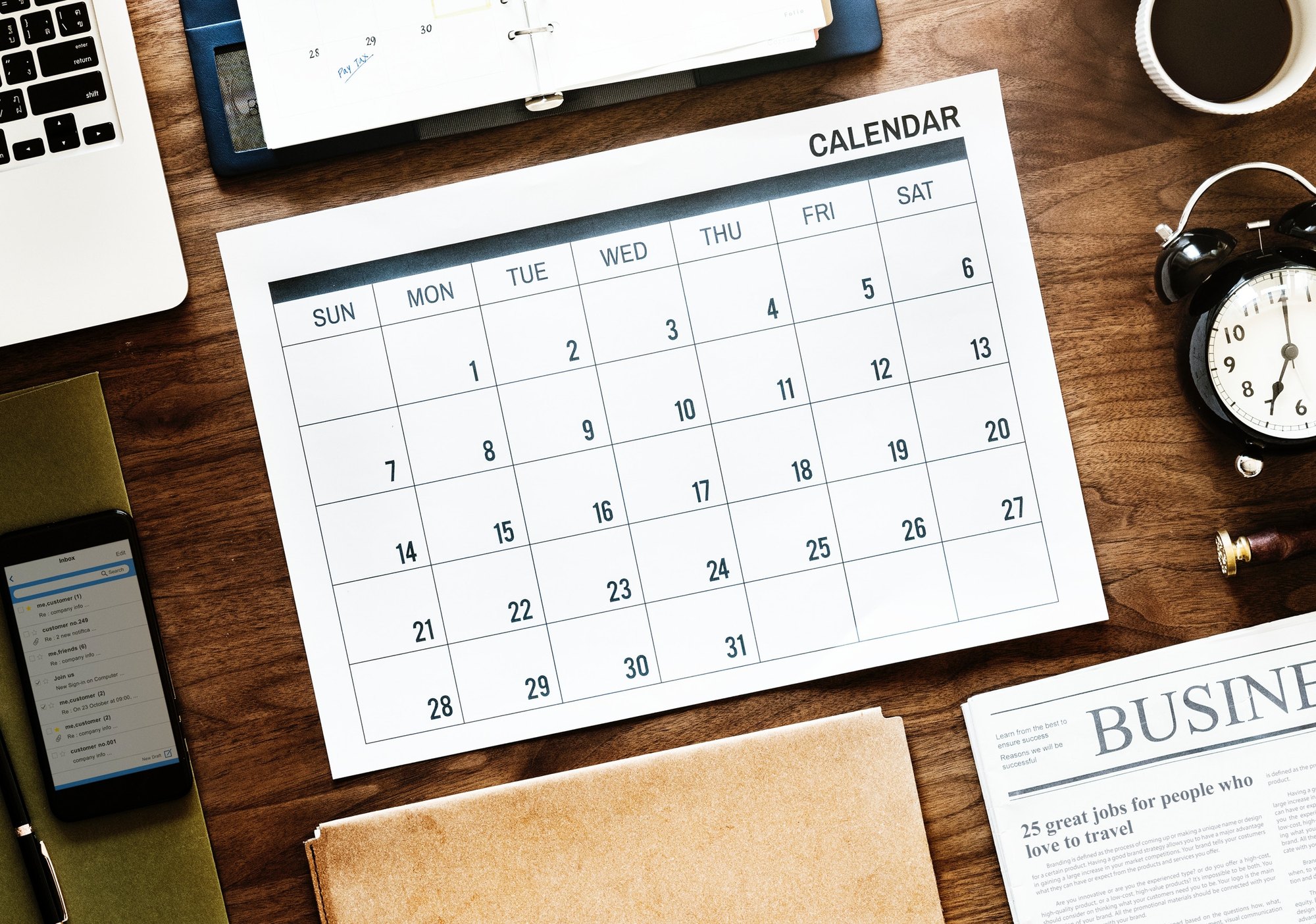 Printed monthly calendar on a wooden desk with a laptop, coffee cup, and smartphone nearby.