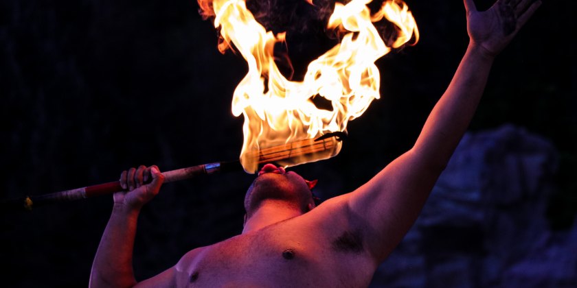 Shirtless fire performer holds a flaming torch near his face against a dark background.