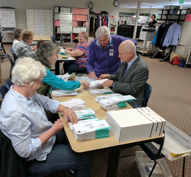 Election workers sorting stacks of mail ballots and envelopes at a table in an elections office.