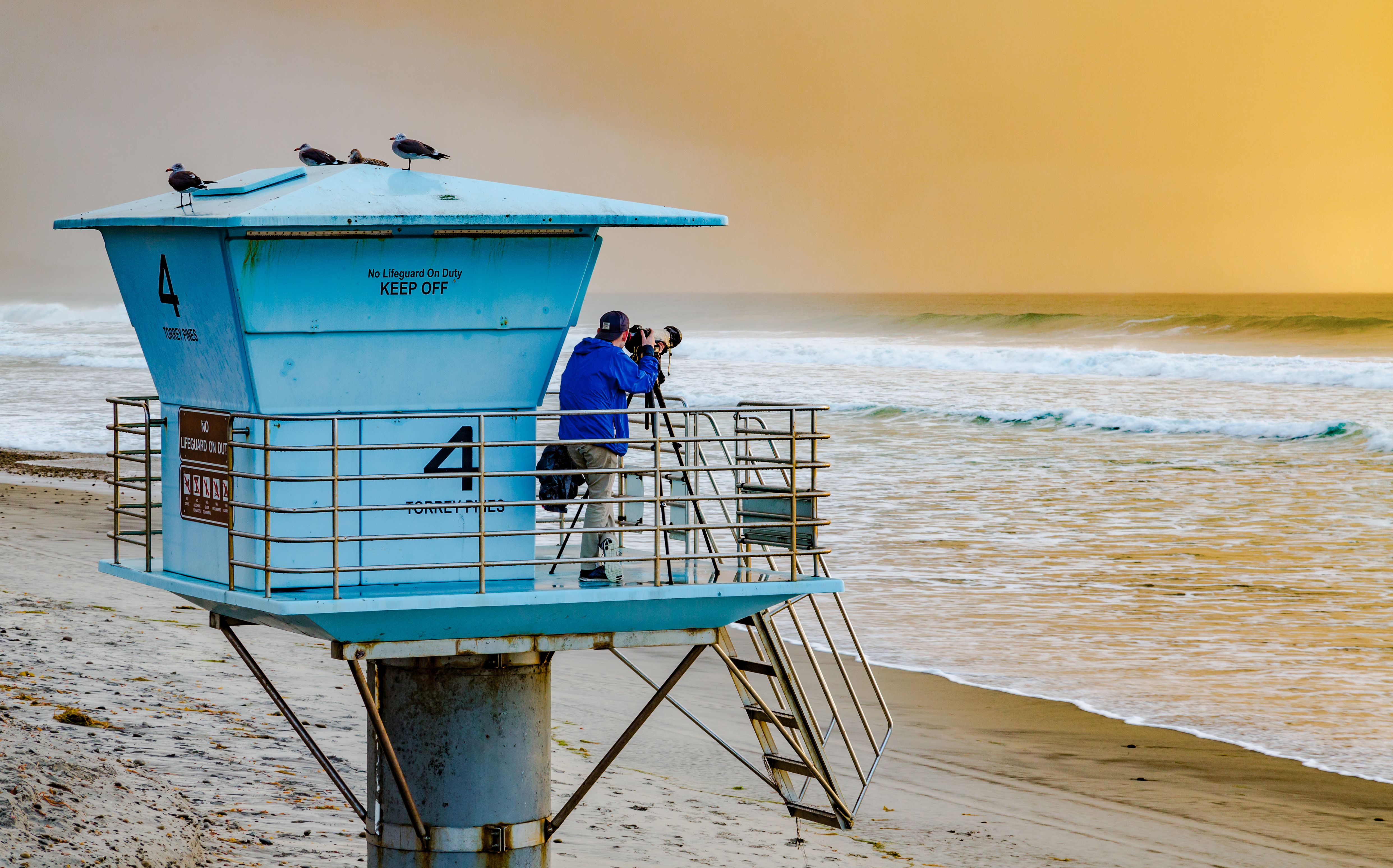Blue lifeguard tower on a sandy beach at sunset, person visible on the observation deck.