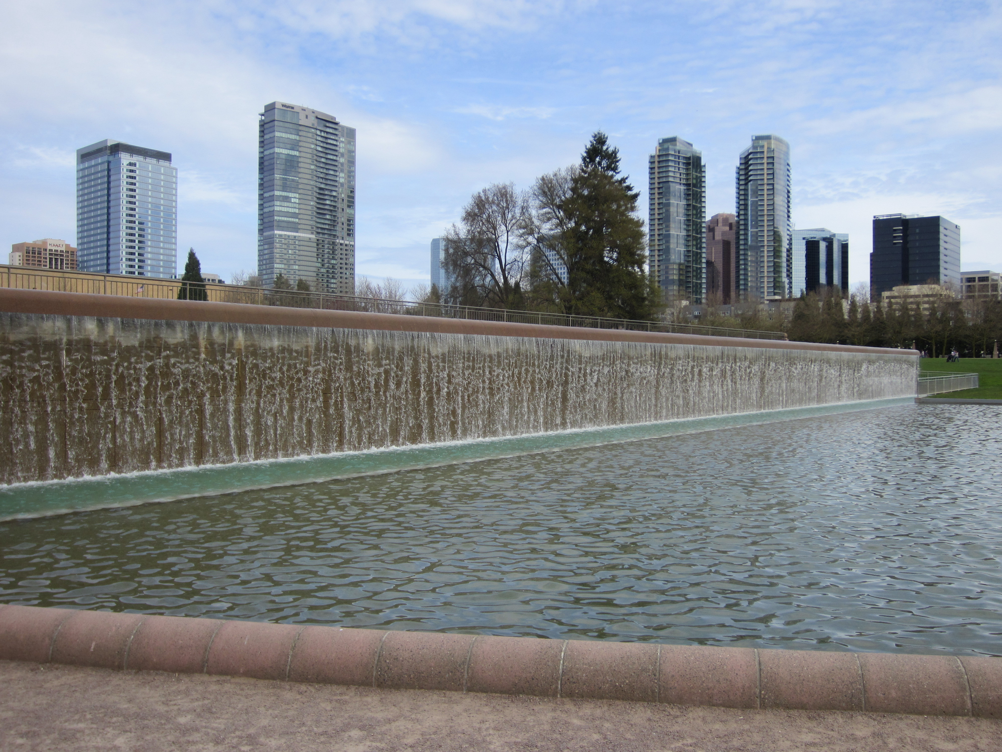 A cascading waterfall flows into a reflecting pool with downtown high-rise buildings visible behind.