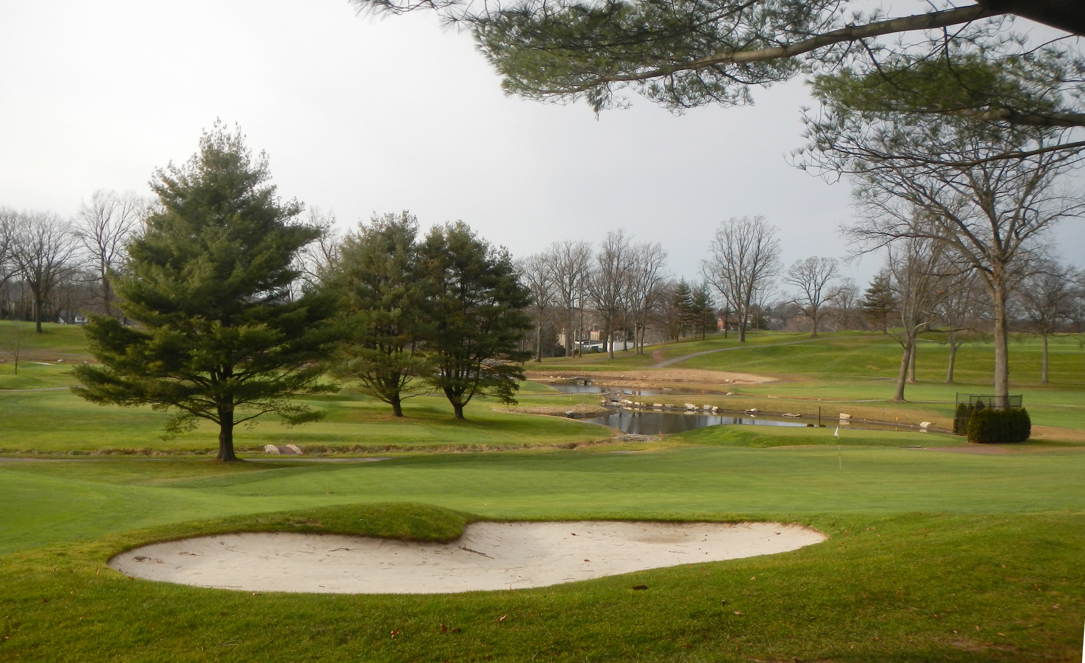 A manicured golf course green with a sand bunker, pond, and trees under an overcast sky.