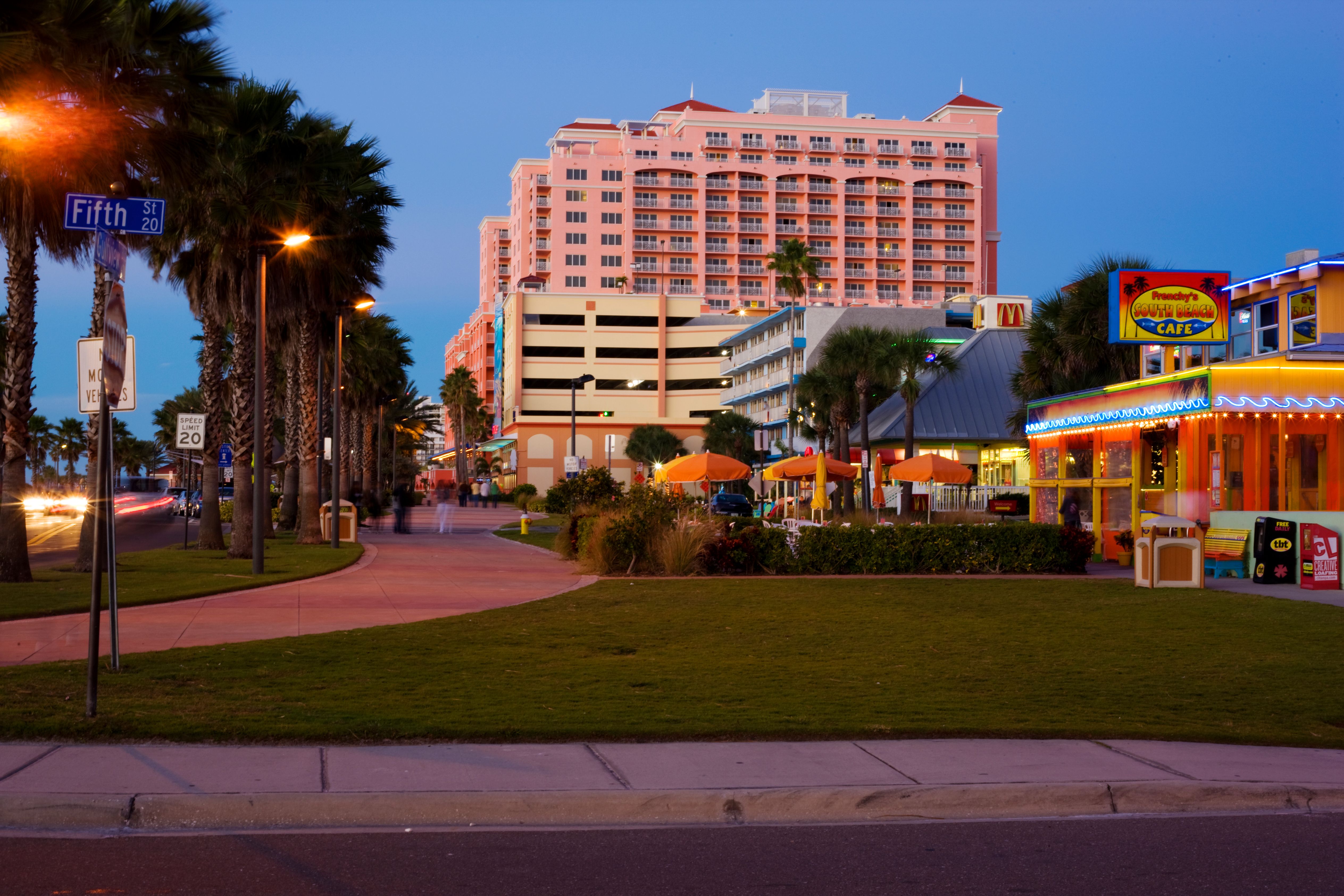 Palm-lined beachside boulevard at dusk with a lit café and large resort hotel.