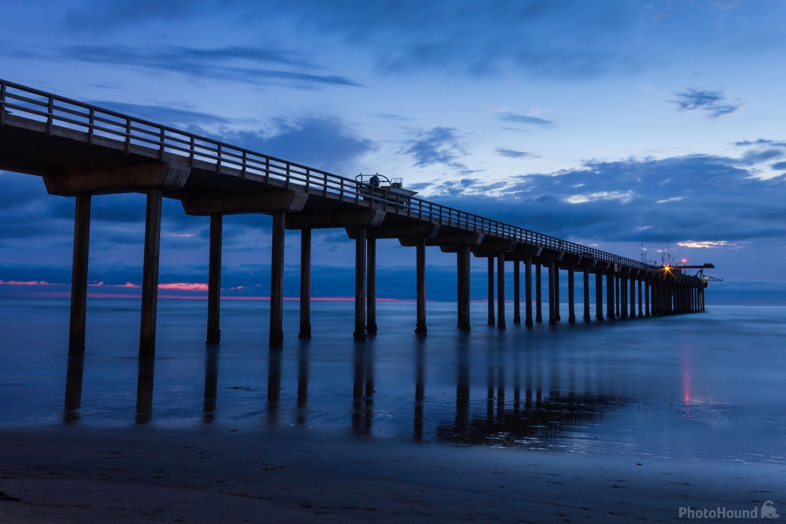 Long pier extending over calm water at twilight with lights visible at its far end.