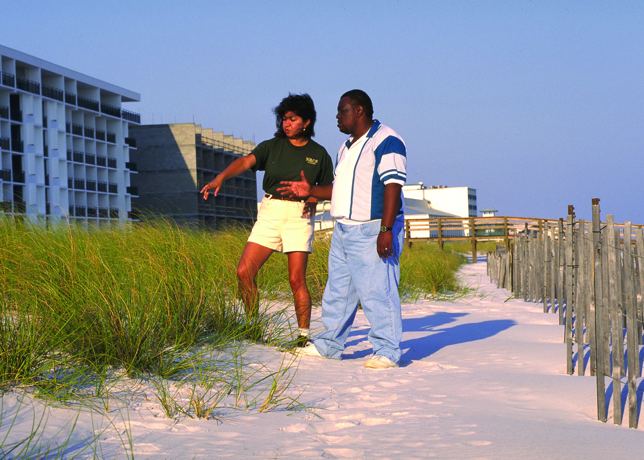 Two people stand beside wooden sand fencing with sea oats growing on a sandy beach dune.