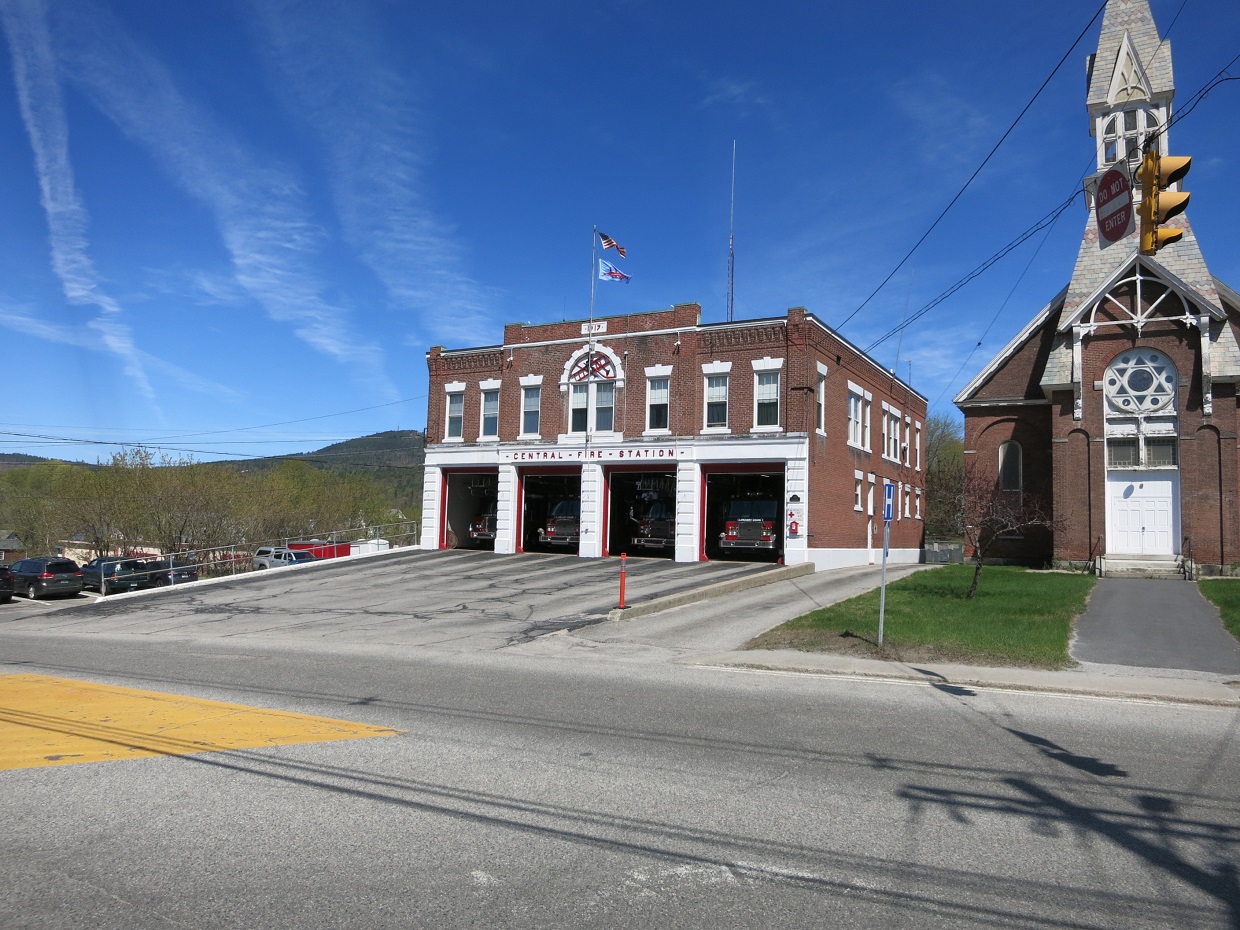 Red brick fire station with three open bays and fire trucks visible inside.