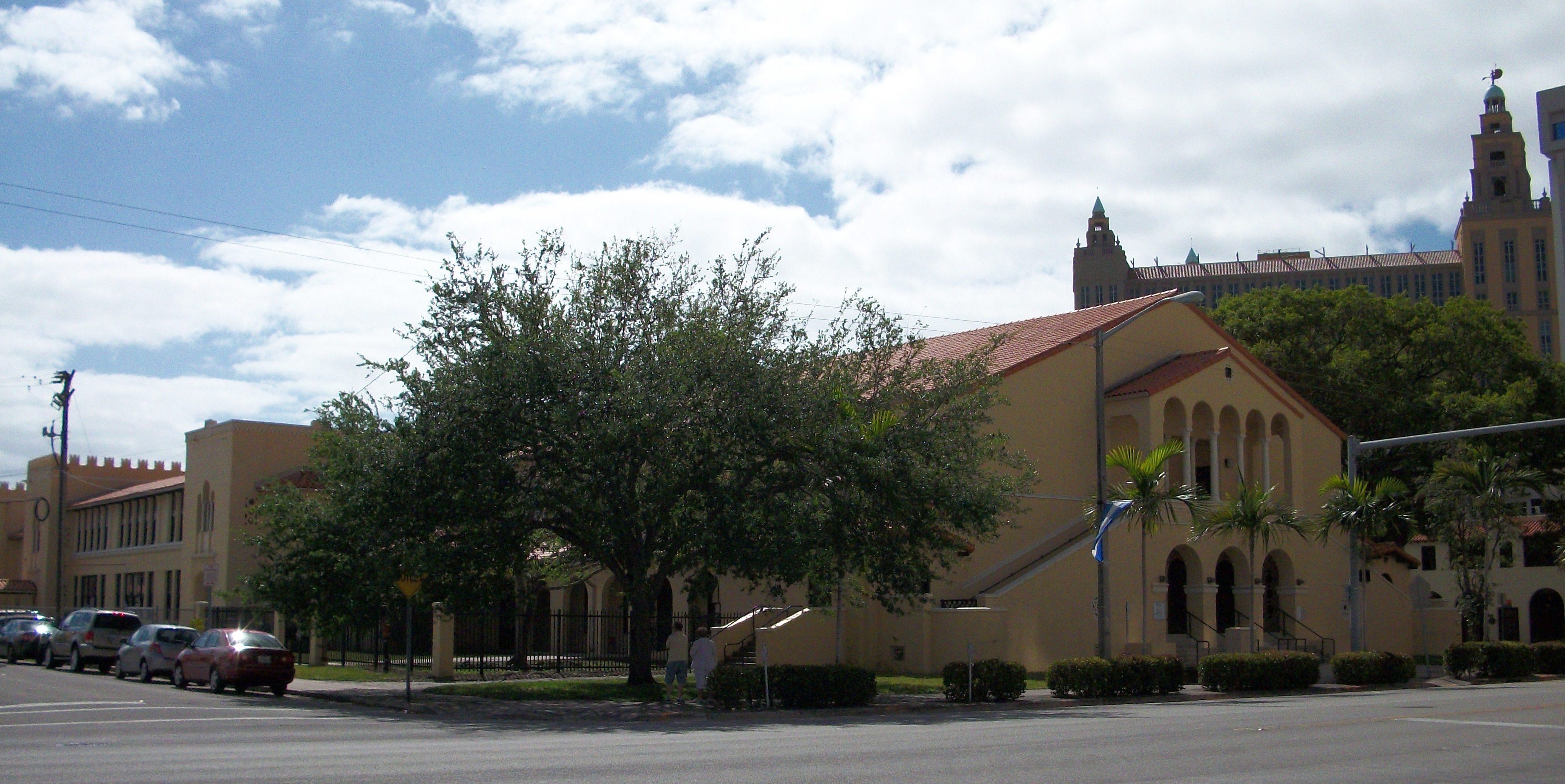 Yellow stucco school building with red tile roof and trees in front.