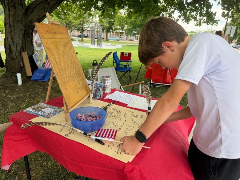 A young person signs a large parchment document at a table with a red tablecloth and patriotic decorations.