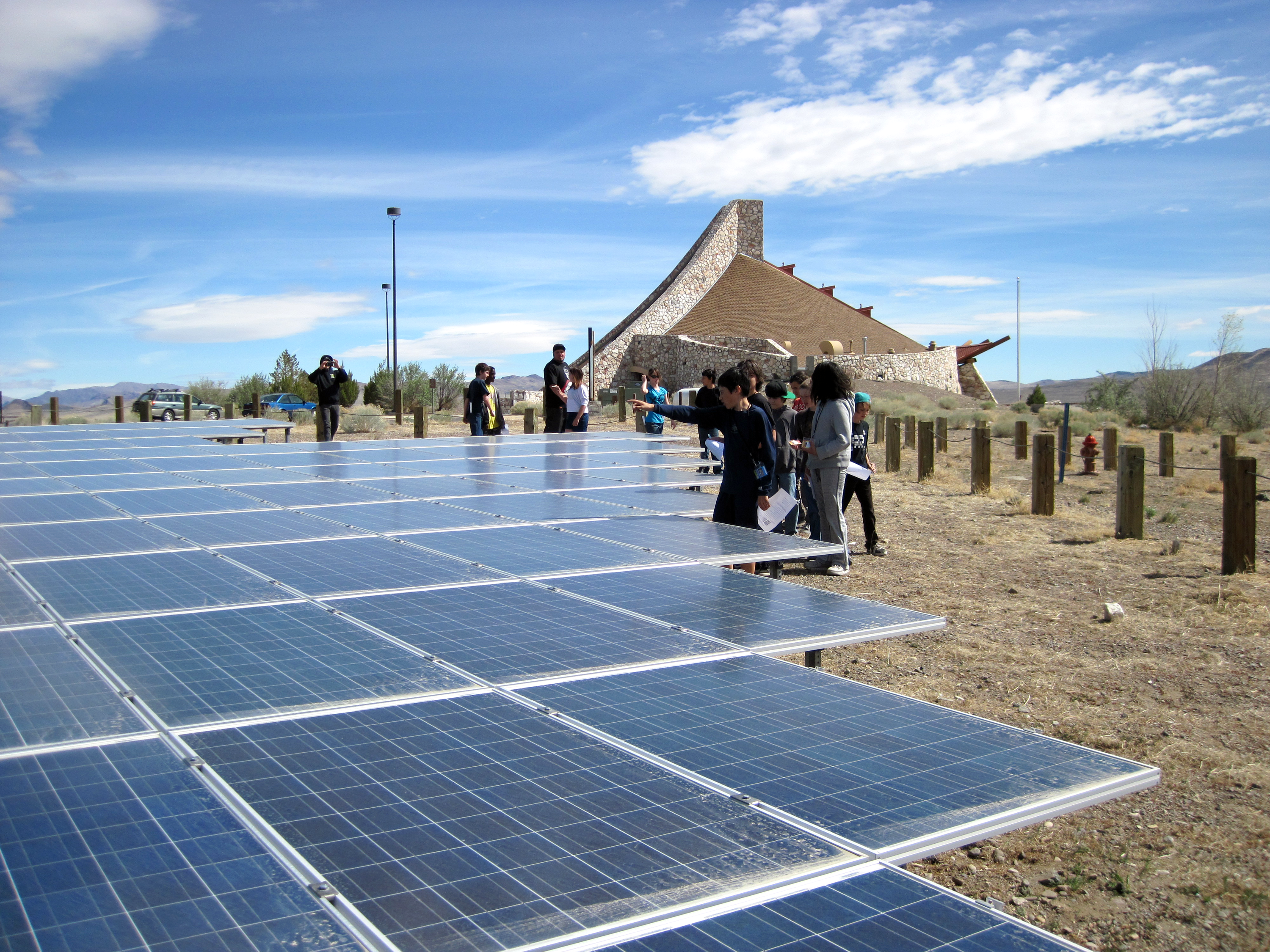Group of children and adults standing beside rows of outdoor solar panels.