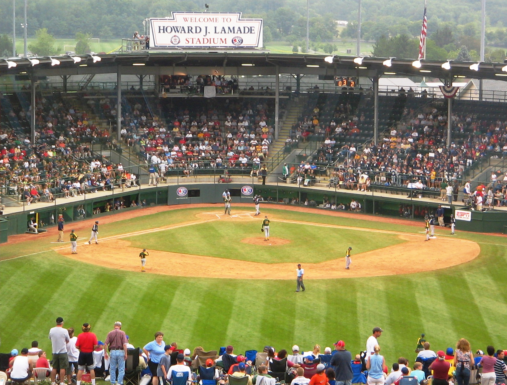 Packed baseball stadium with green wooded hills visible beyond the outfield.