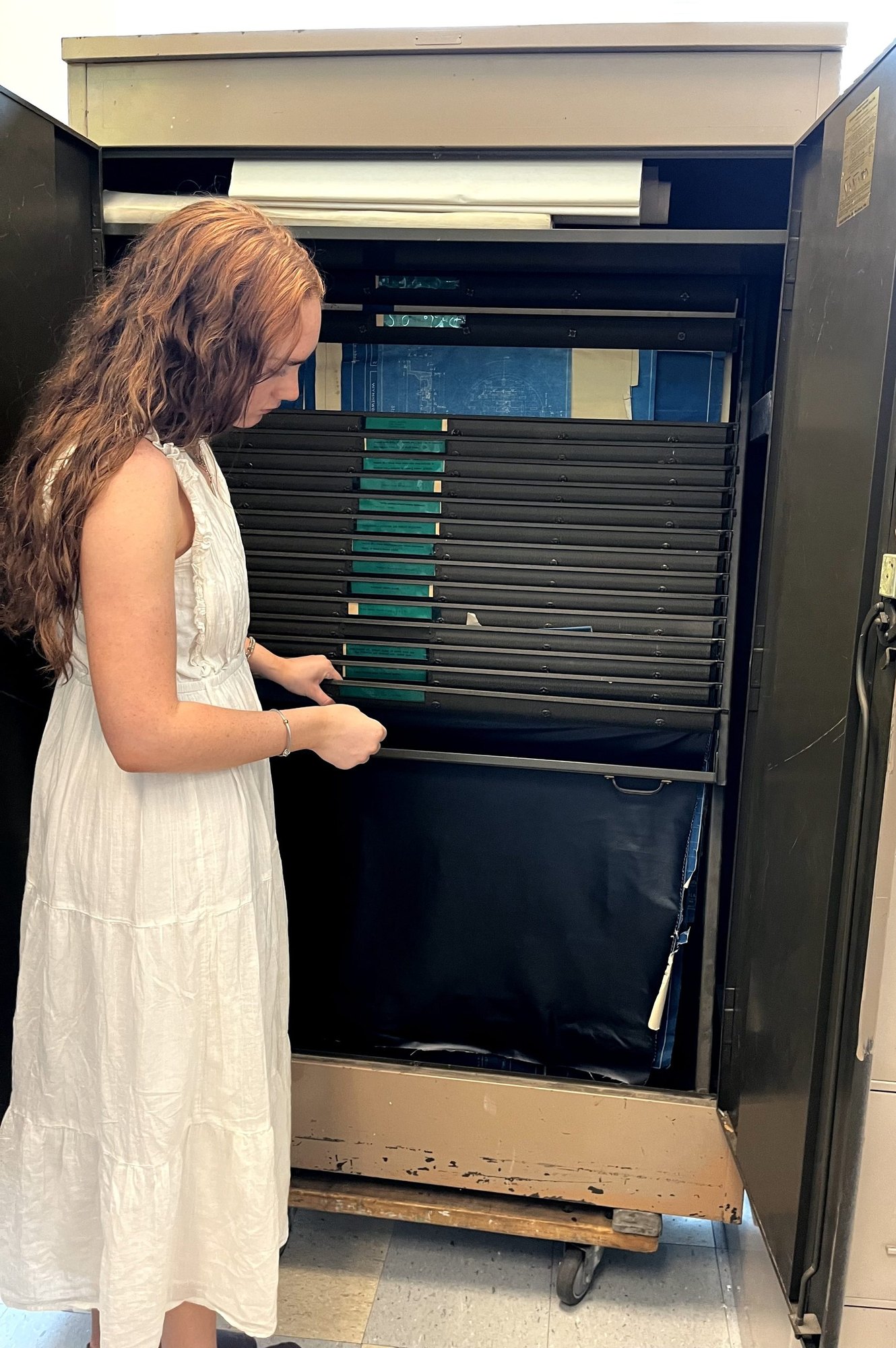 A young woman in a white dress looks into an open flat-file archival storage cabinet.