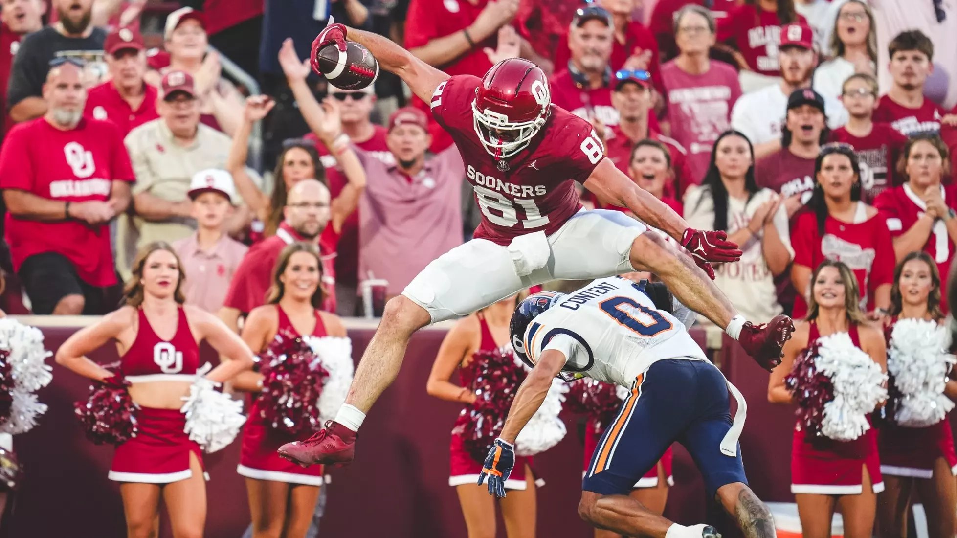 A football player in a red uniform leaps over a defender near the end zone.