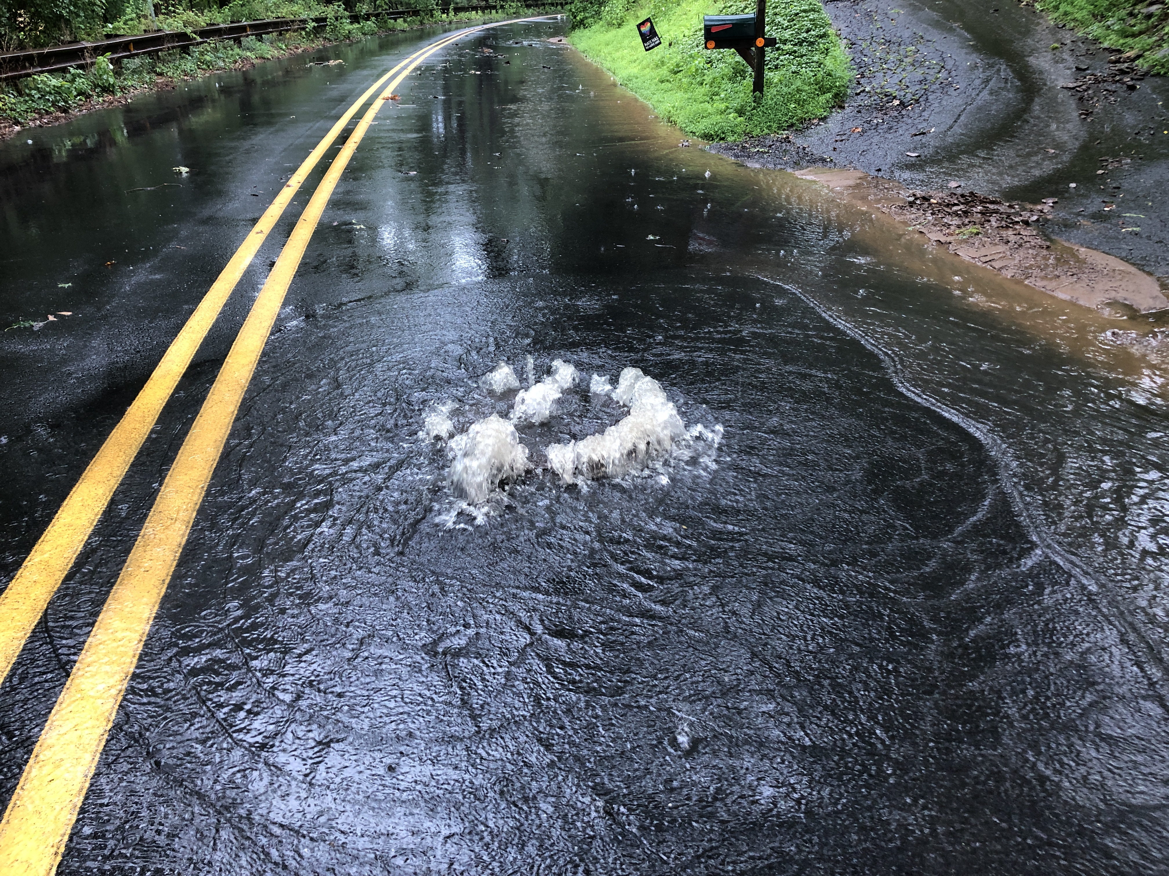 Floodwater erupts upward from a manhole cover on a water-covered two-lane road.