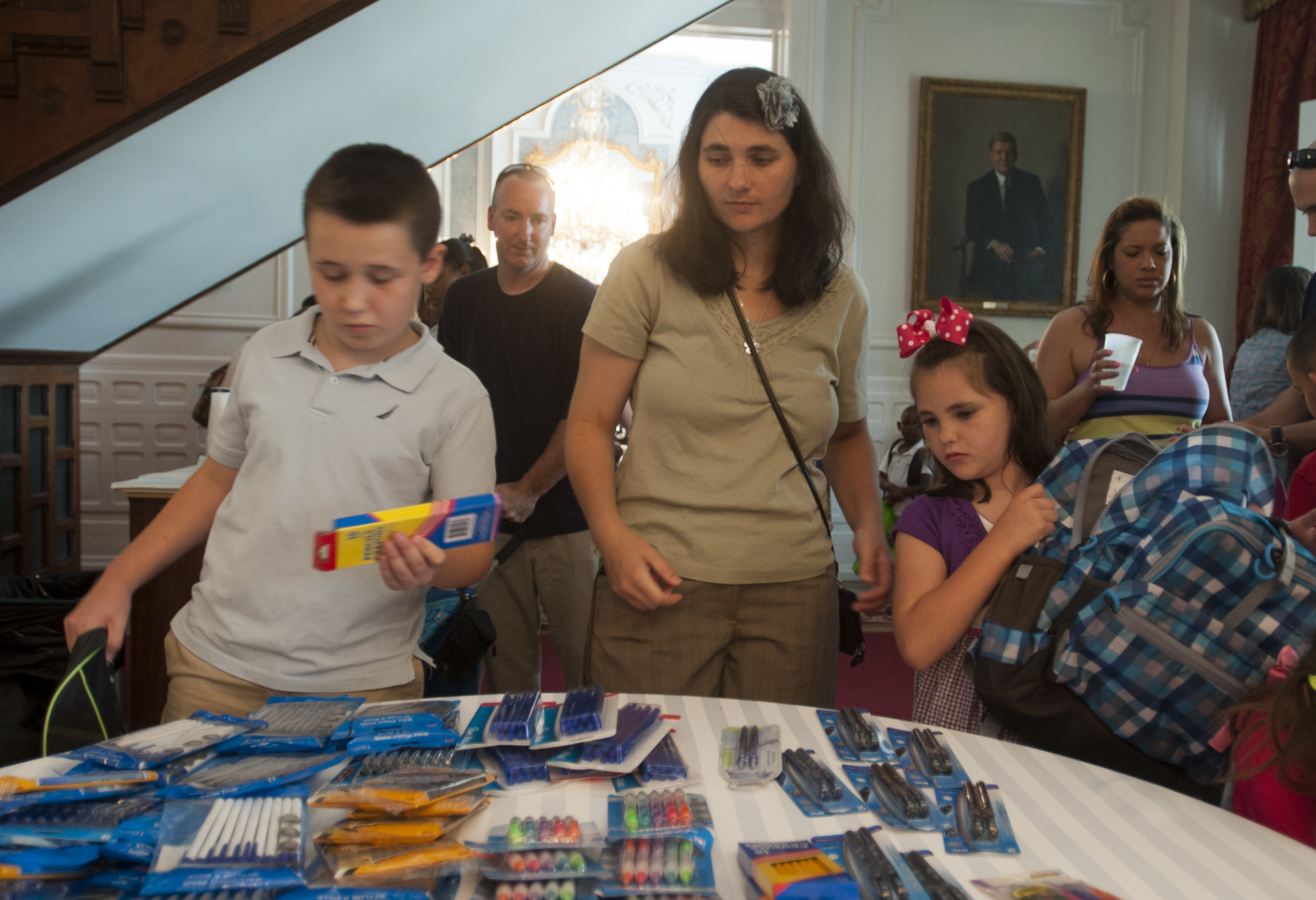A woman and two children select school supplies from a table at an indoor event.