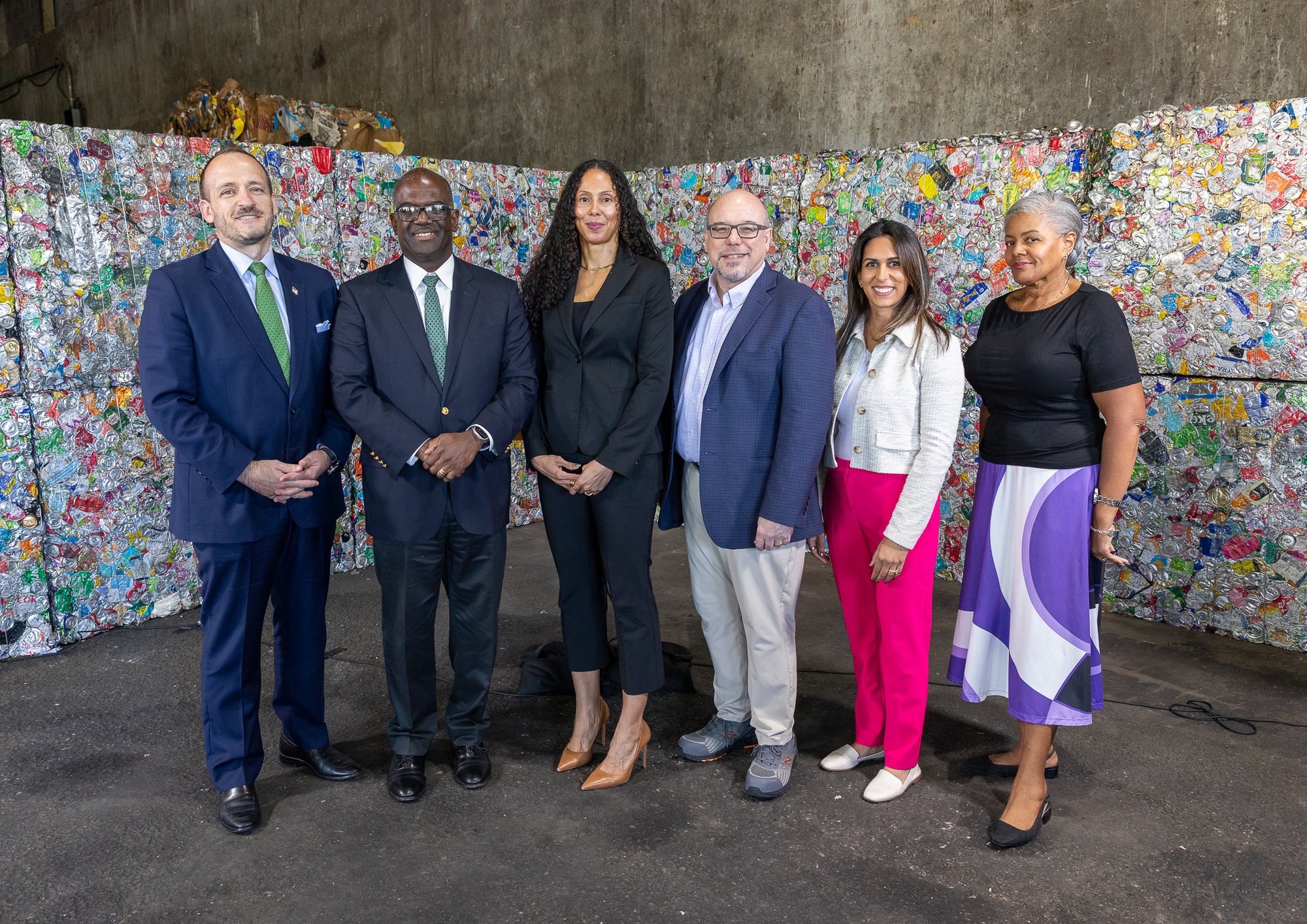 Six officials pose in front of large bales of crushed aluminum cans inside a recycling facility.