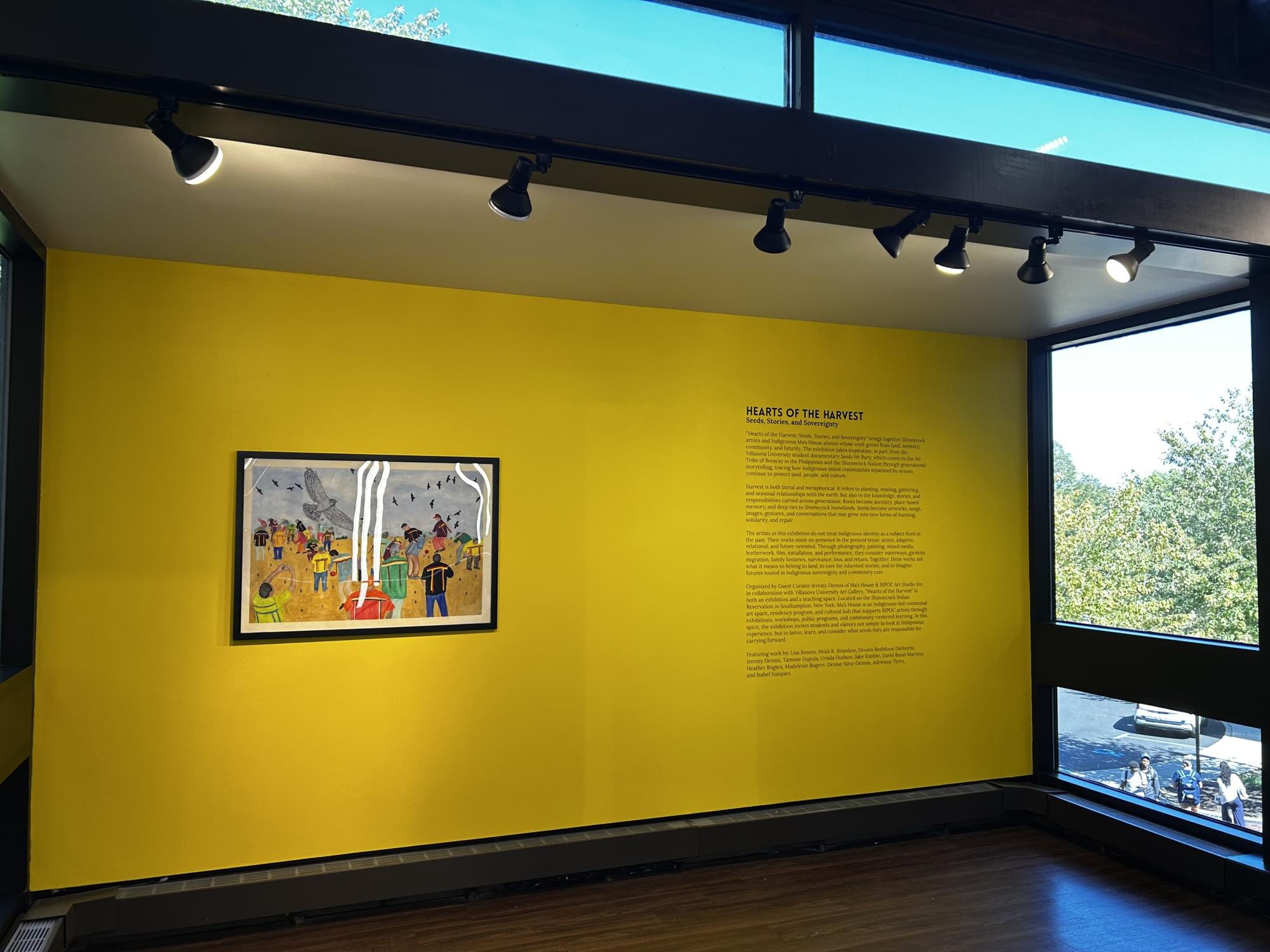 Yellow gallery wall with a framed artwork and an exhibition title panel under track lighting.