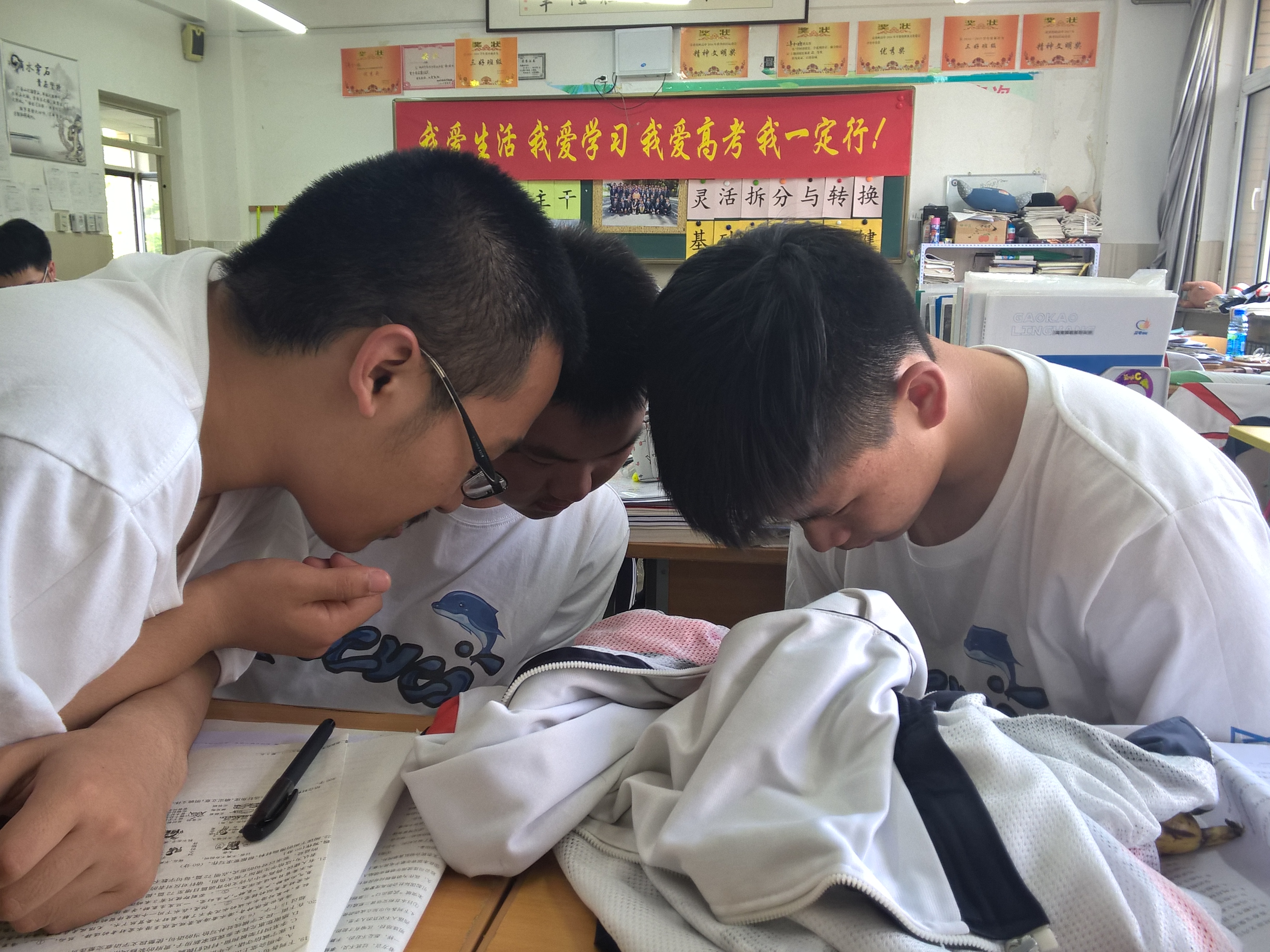 Three students in white shirts huddle over a desk in a classroom.