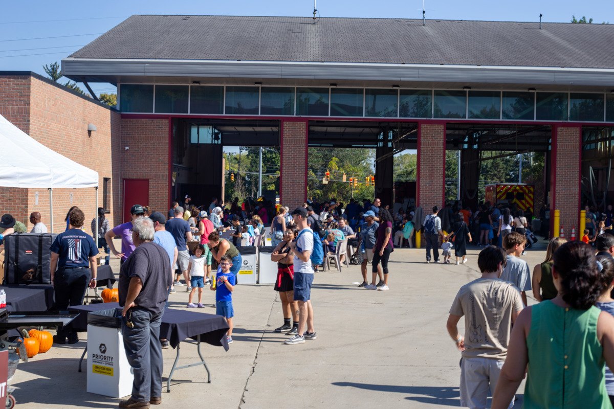 A large crowd of adults and children gathered outside a brick building with red-trimmed bay doors on a sunny day.