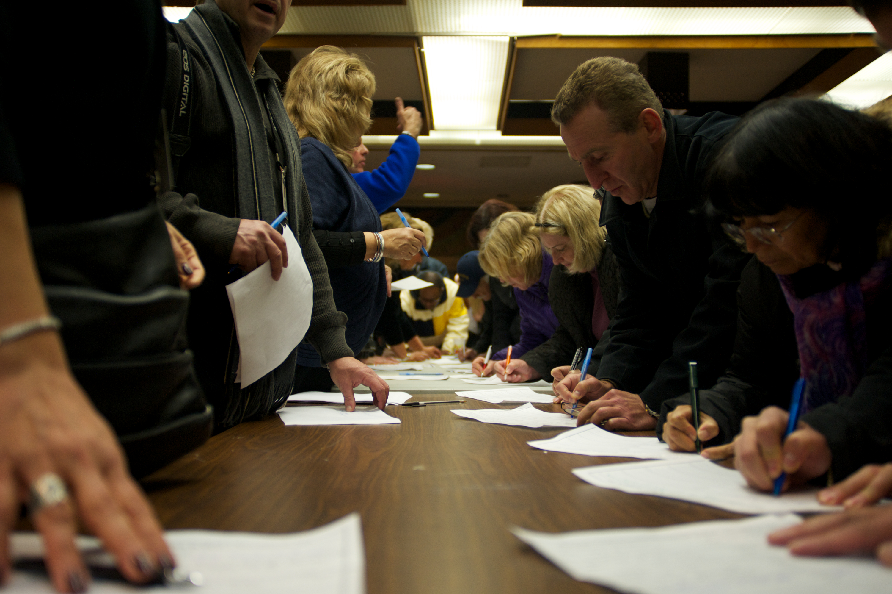People standing at a table writing on paper at an indoor community meeting.