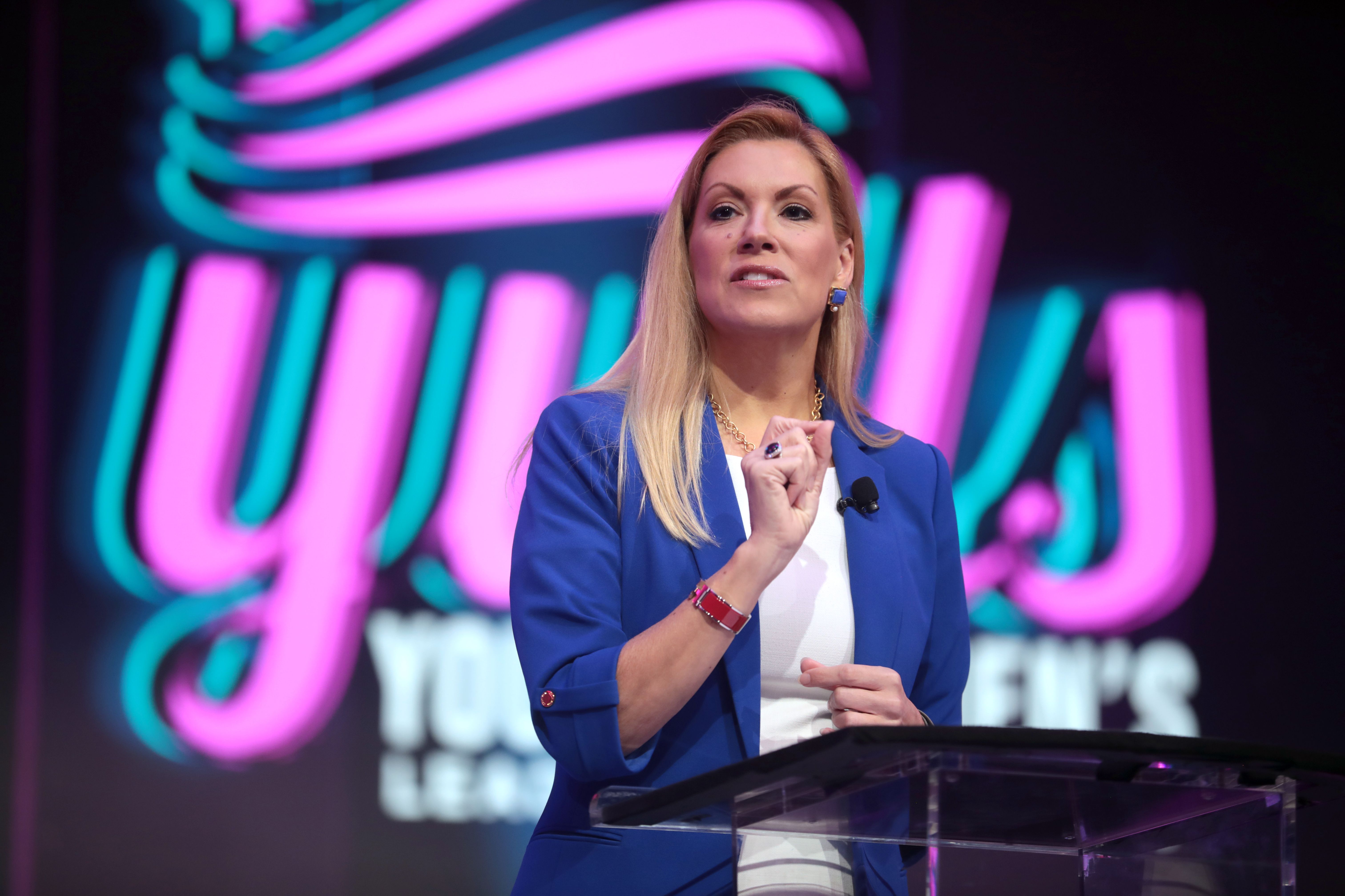Woman in blue blazer speaking at a clear podium with a neon leadership summit sign behind her.
