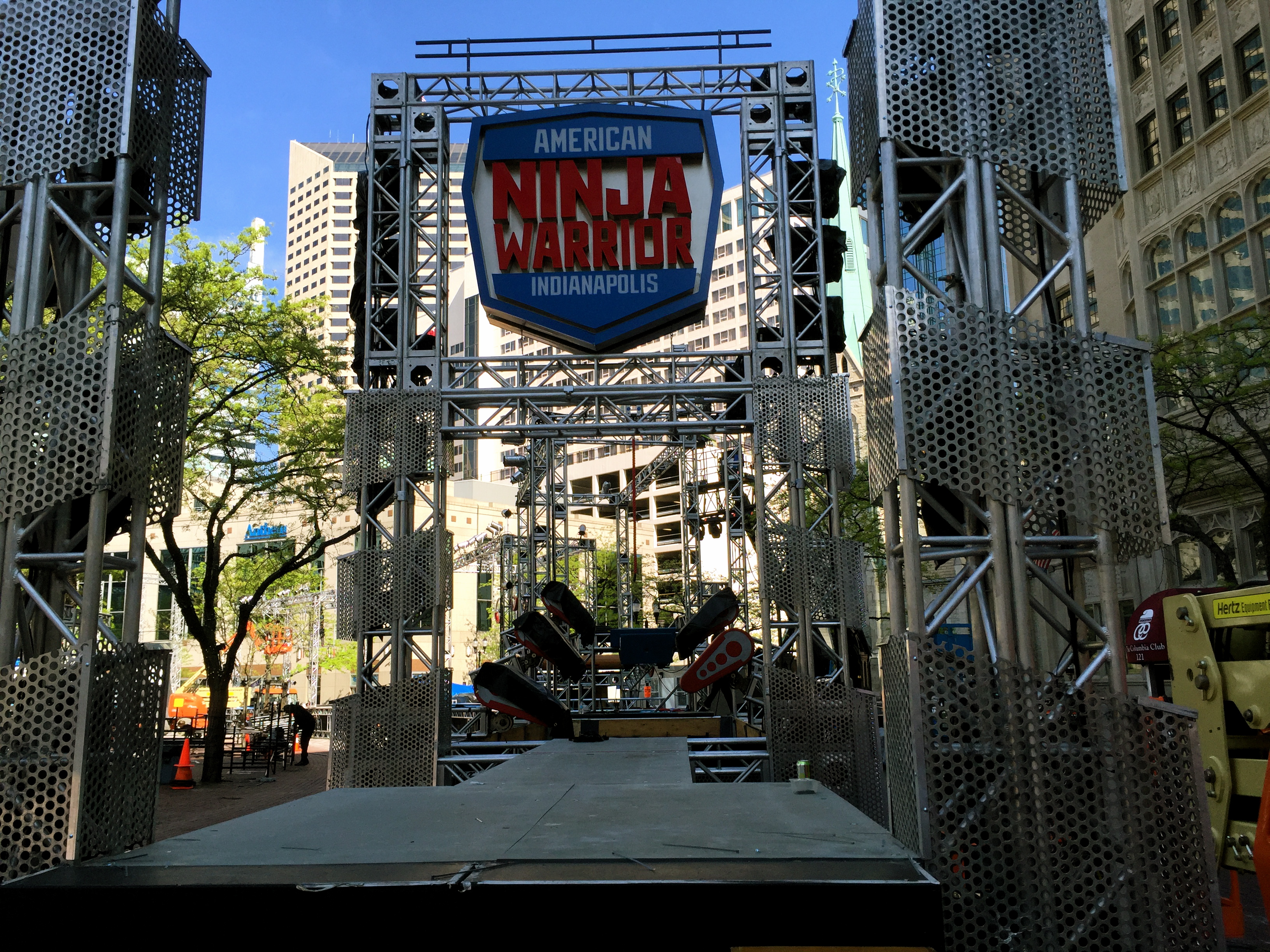 Metal entrance structure with American Ninja Warrior sign and scaffolding at an outdoor venue.