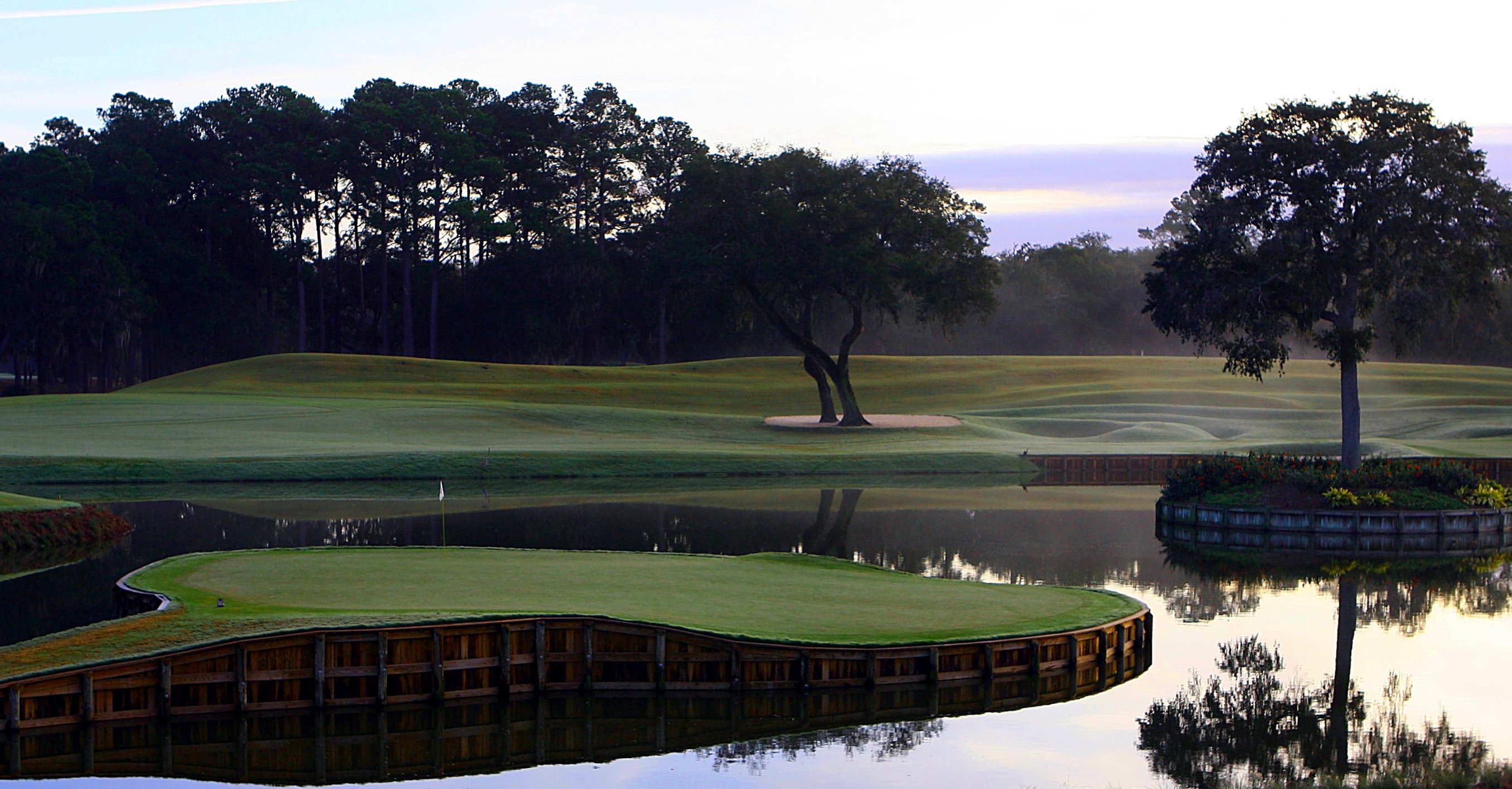 Island green 17th hole at TPC Sawgrass surrounded by calm water at dusk.