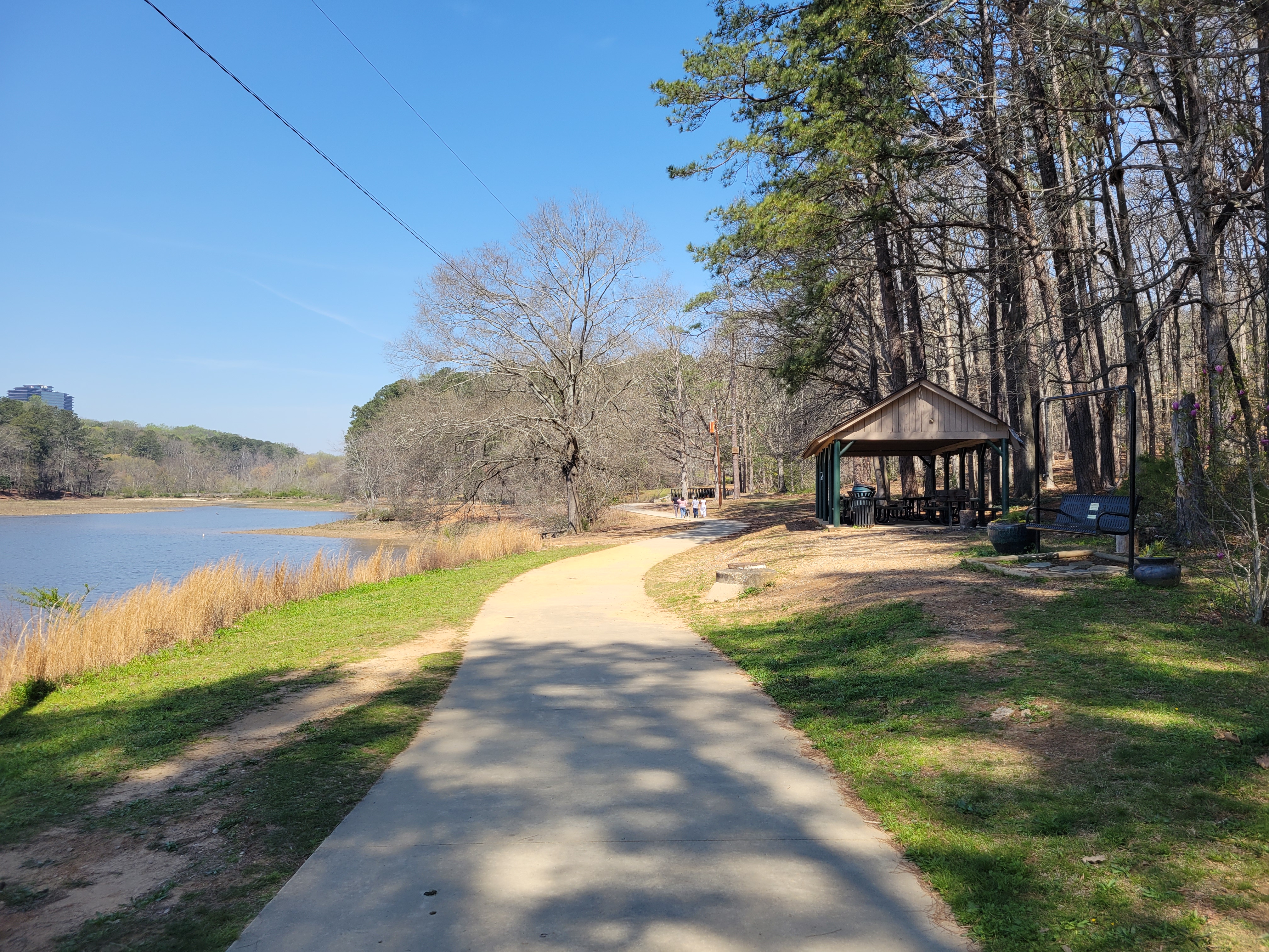 Paved path alongside a lake with a wooden pavilion and tall pine trees.