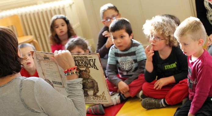An adult holds open a picture book while several young children sit on the floor listening.
