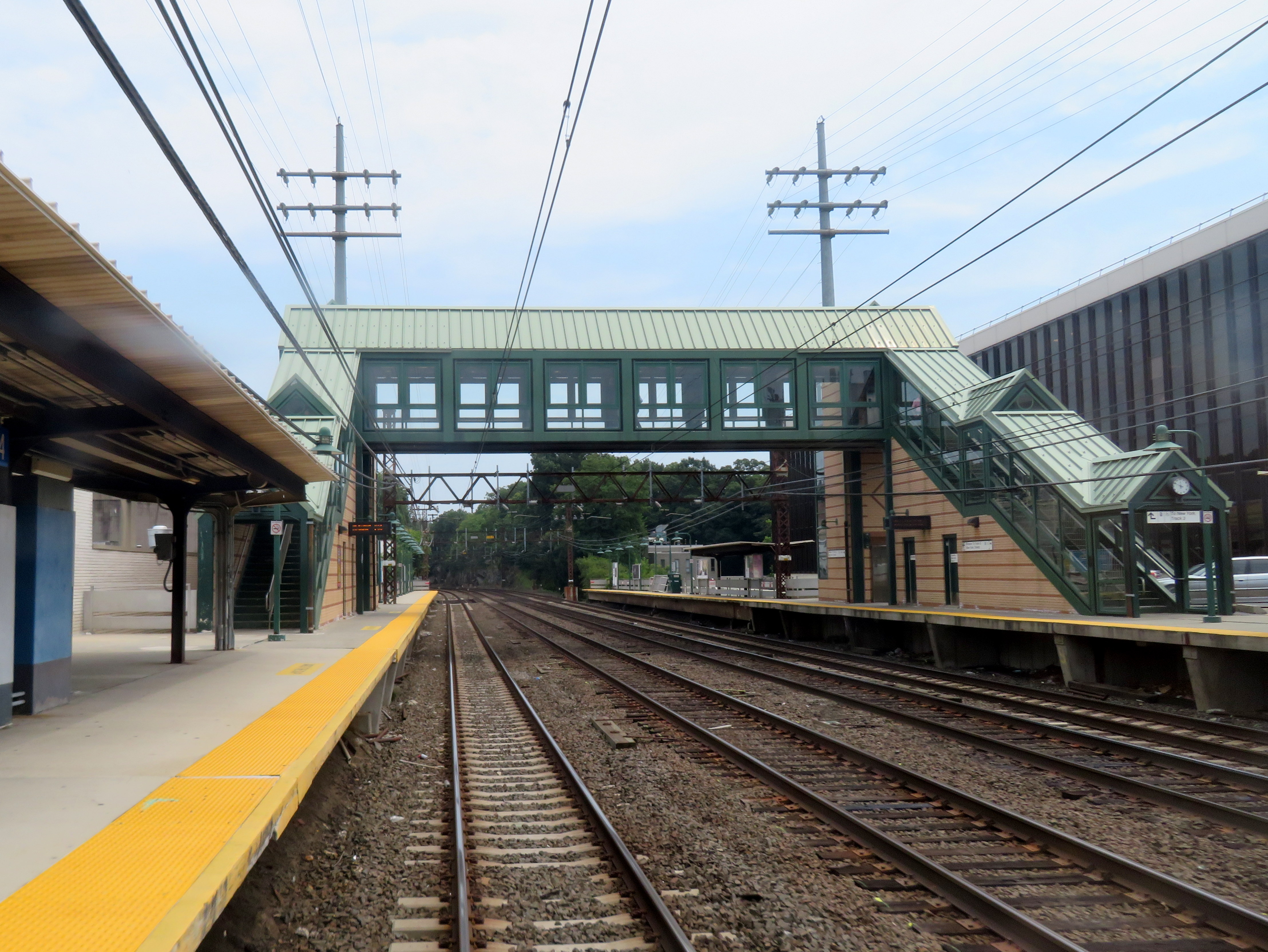 Commuter rail station with two platforms, pedestrian overpass bridge, and overhead catenary wires.