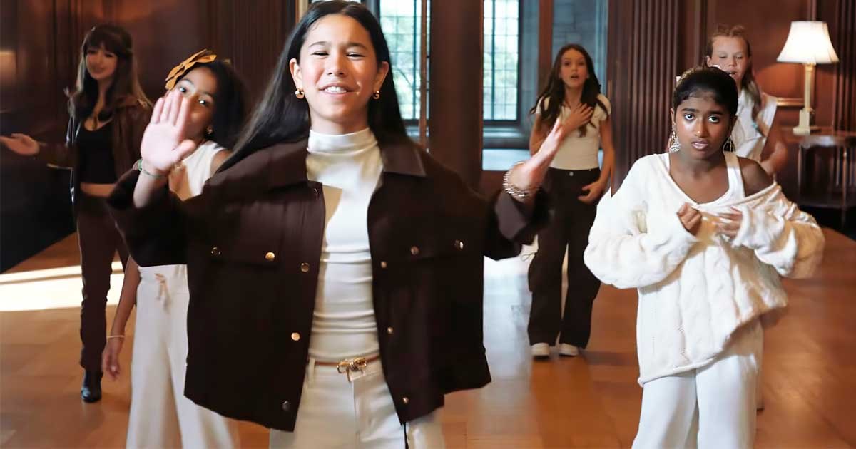 Young female performers in white and brown outfits dancing in an elegant wood-paneled room.