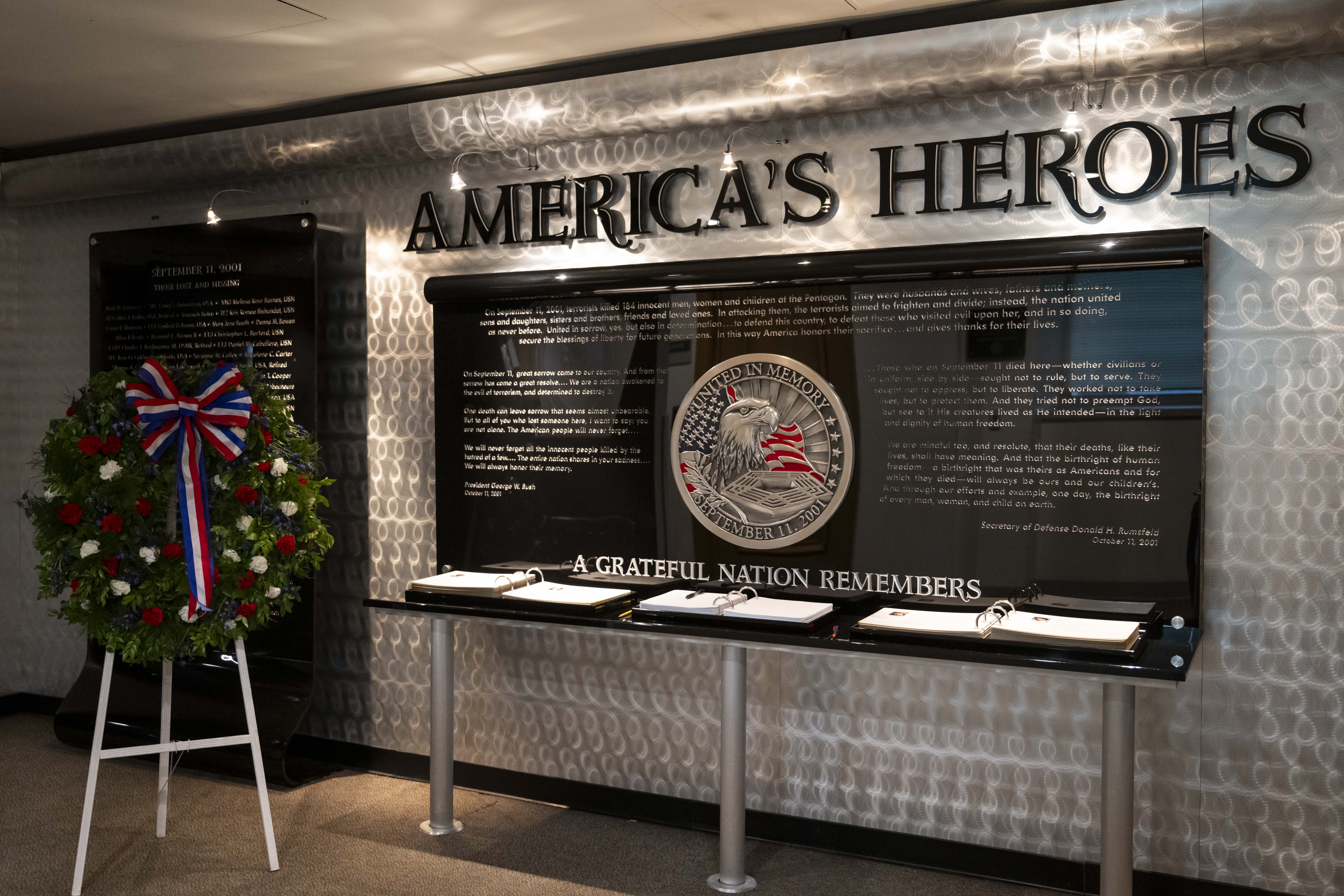 Indoor memorial display with patriotic signage, open books of remembrance, and a red-white-and-blue floral wreath.