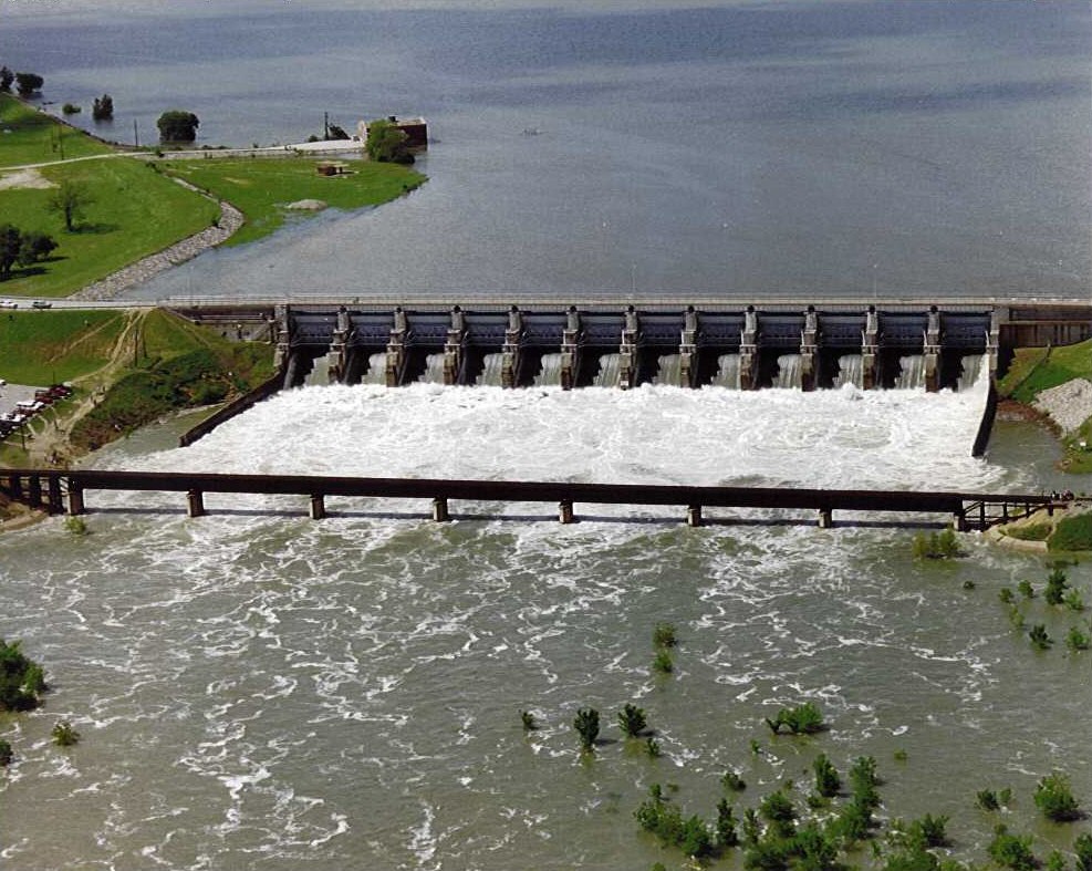 Aerial view of a dam with open spillway gates and a large reservoir behind it.