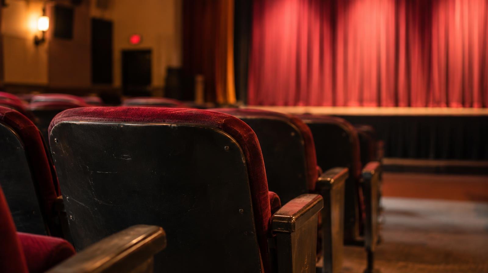 Empty red theater seats face a warmly lit stage curtain in a small playhouse.