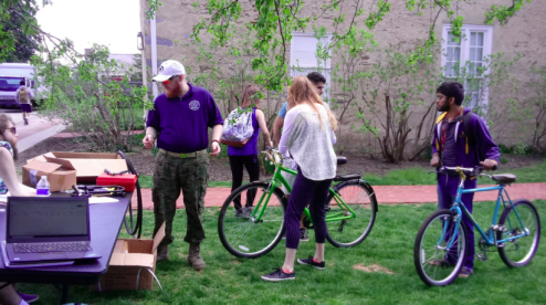 People examine green and teal bicycles on a grassy college campus lawn.