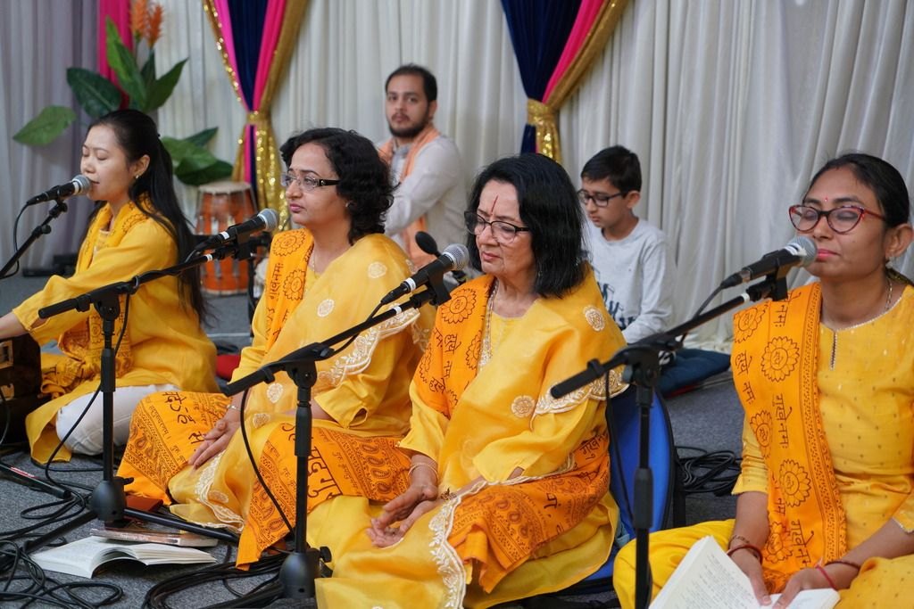 Four women in matching yellow attire sing into microphones at an indoor devotional gathering.