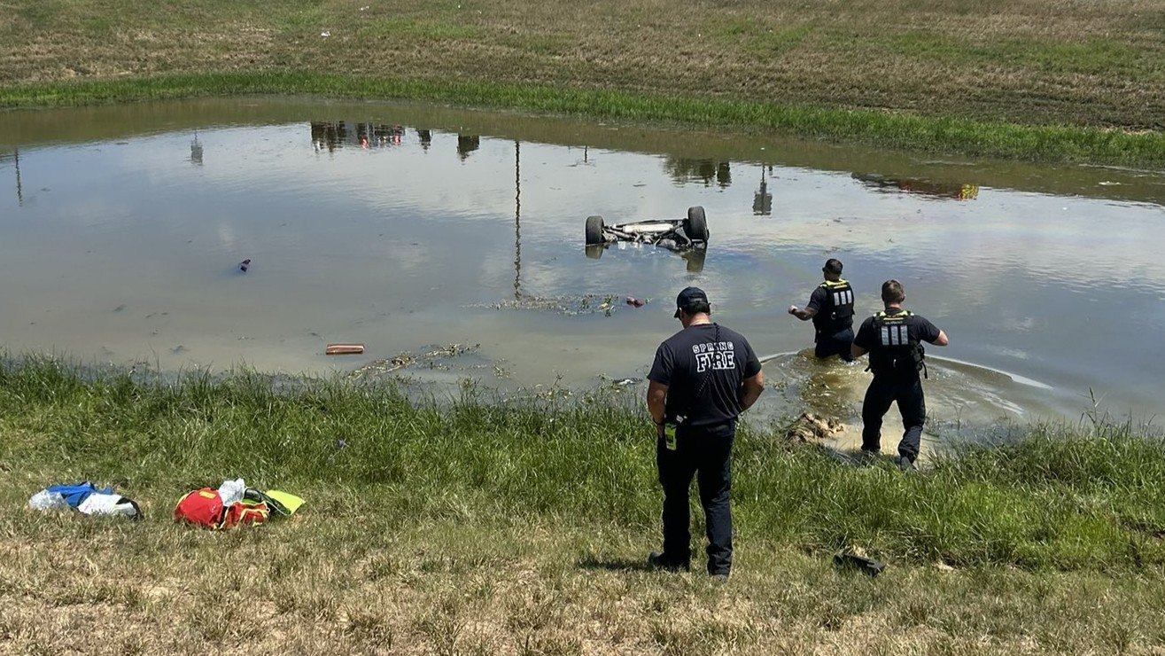 Overturned SUV with wheels up in a murky retention pond, three uniformed responders at the bank.