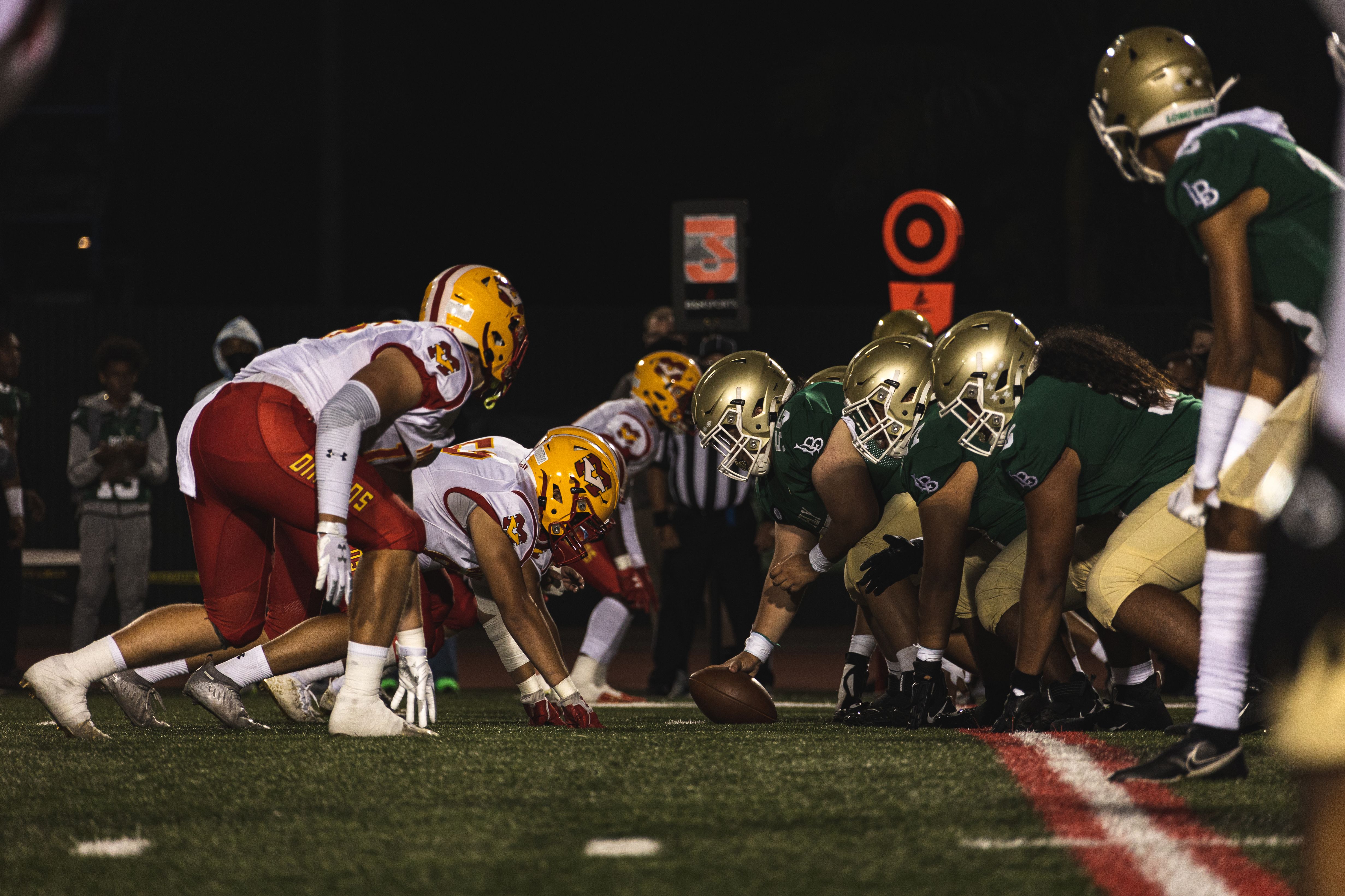 Two high school football teams lined up at the line of scrimmage under stadium lights at night.