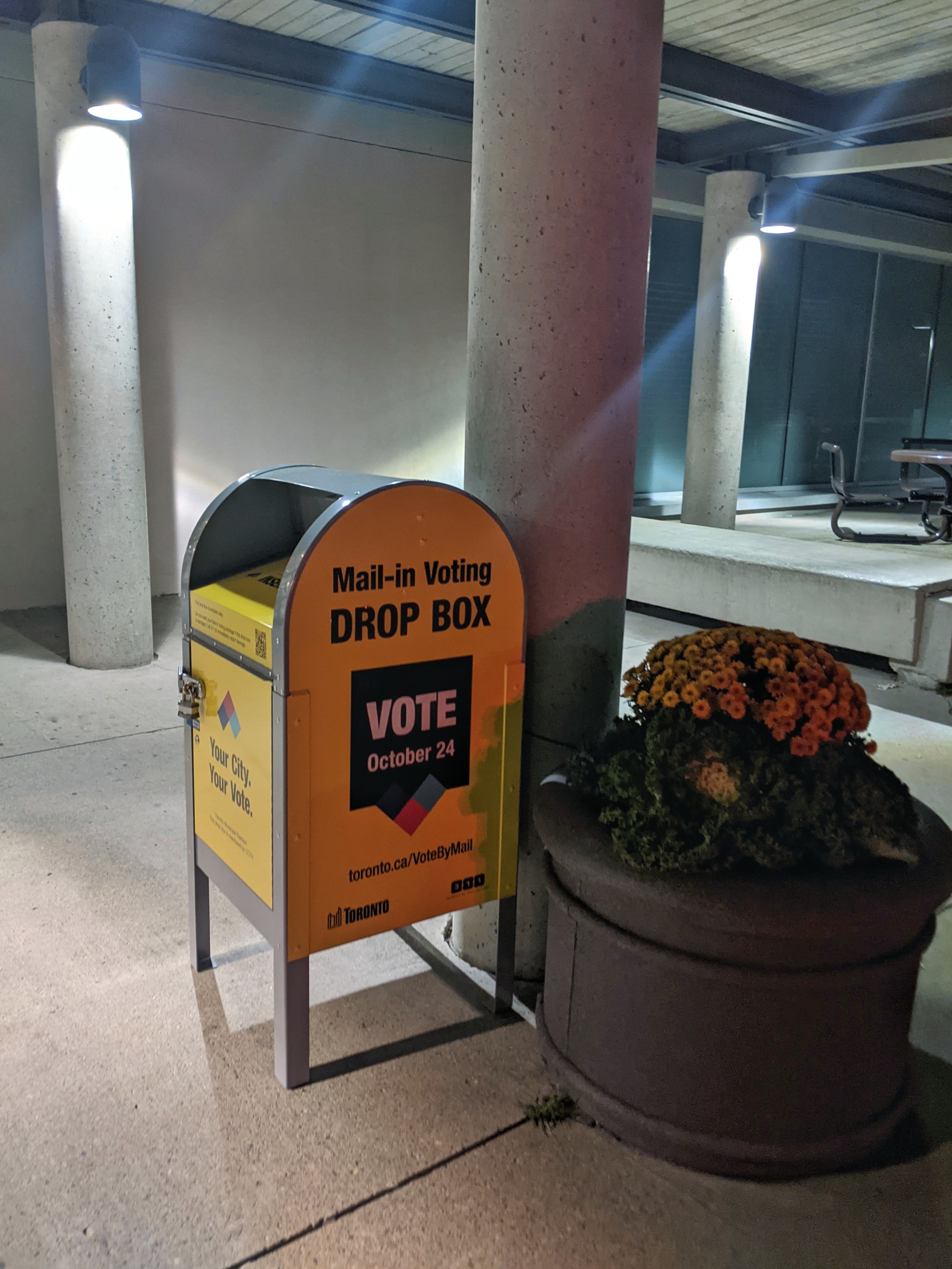 An orange mail-in voting drop box under a covered outdoor area next to a concrete pillar.