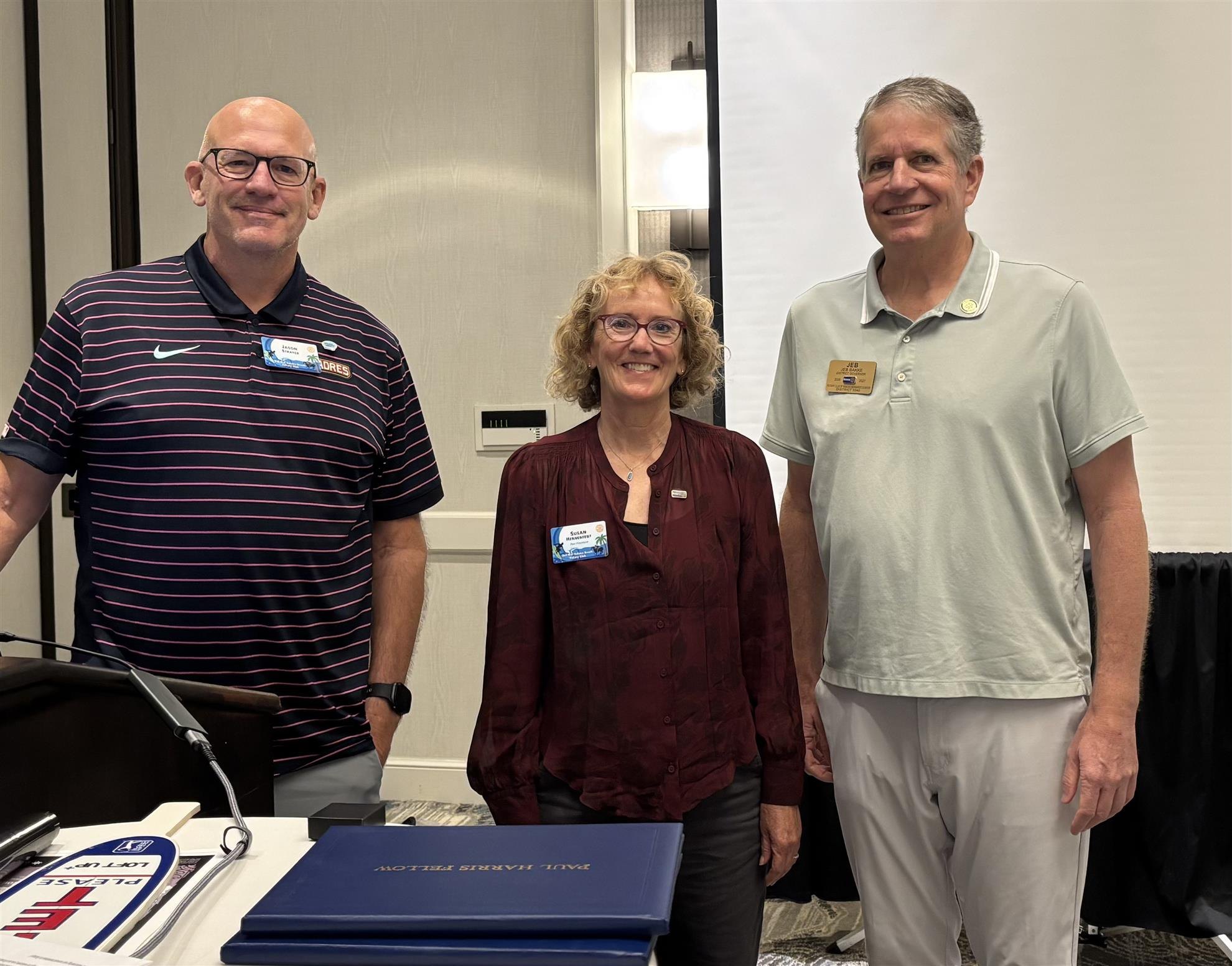 Three smiling adults in name badges stand together in a hotel meeting room with Rotary materials on the table.