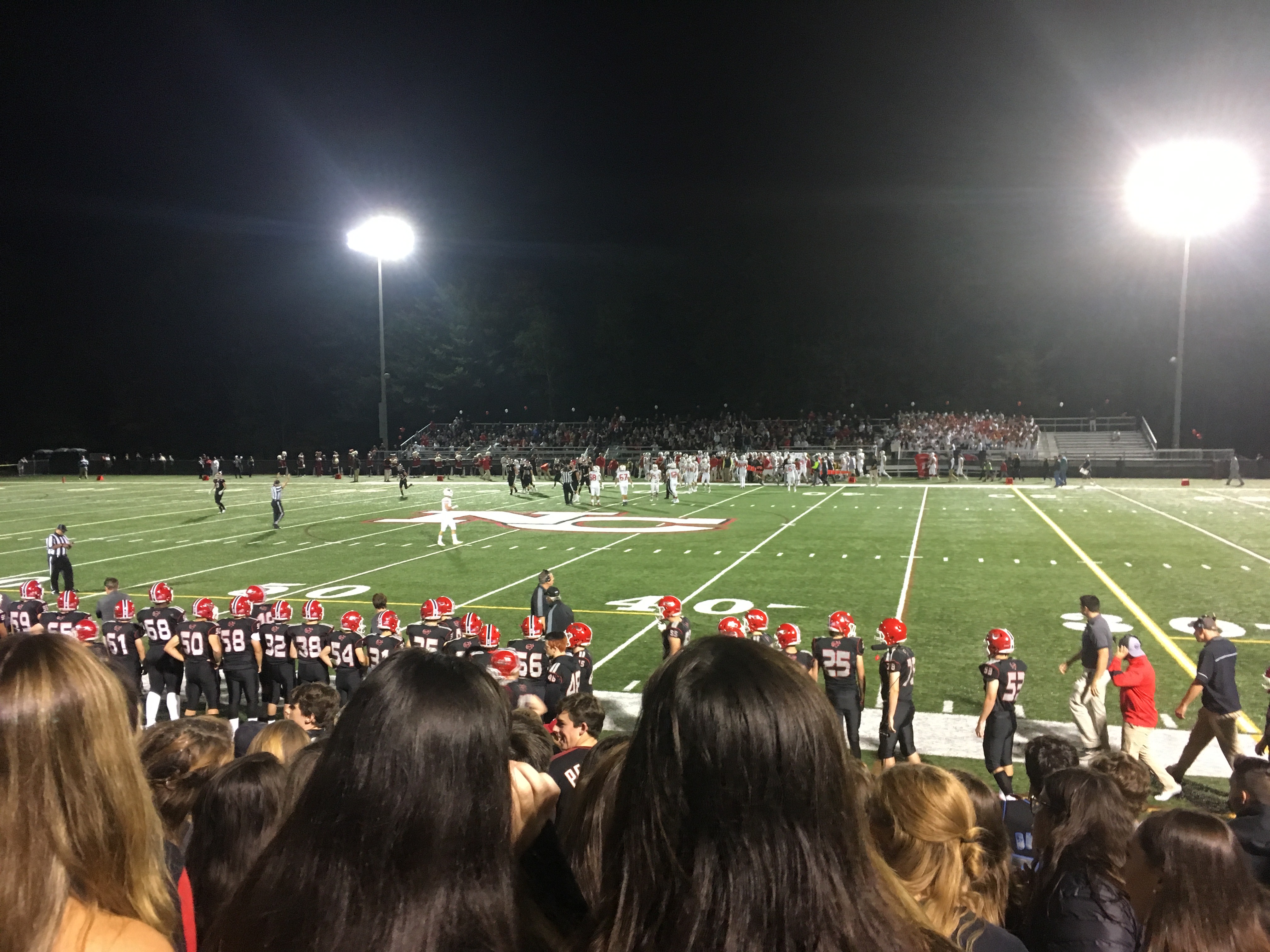 Nighttime high school football game viewed from the student section under stadium lights.