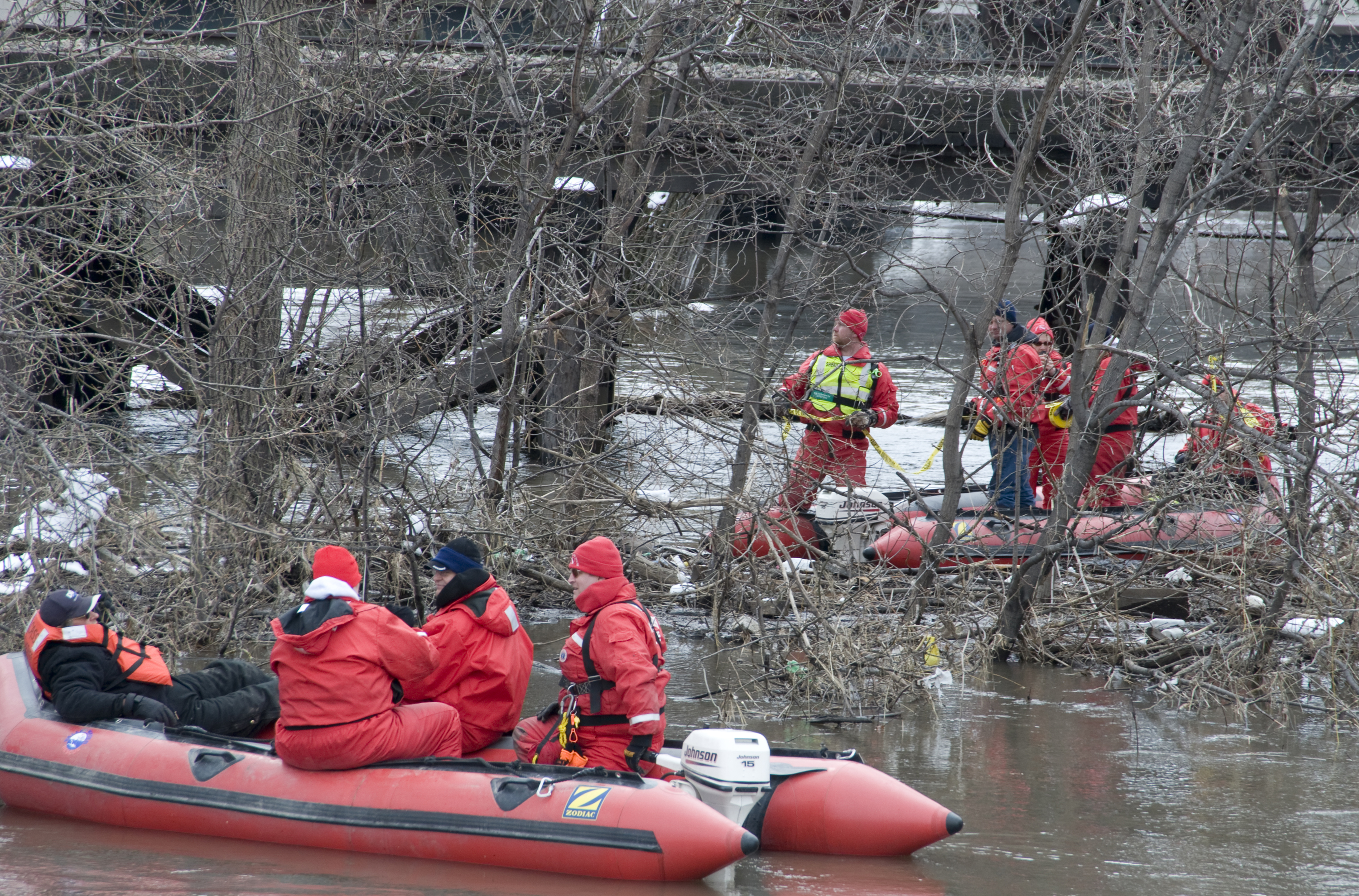 Rescue workers in red dry suits operating inflatable boats on brown floodwaters.