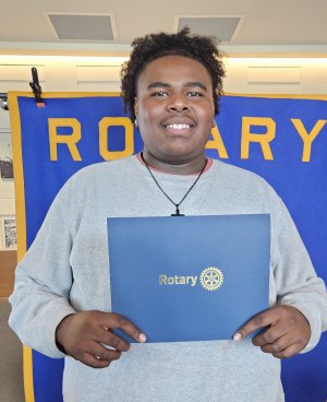 A smiling young man in a gray sweatshirt holds a blue Rotary Club certificate folder in front of a Rotary banner.