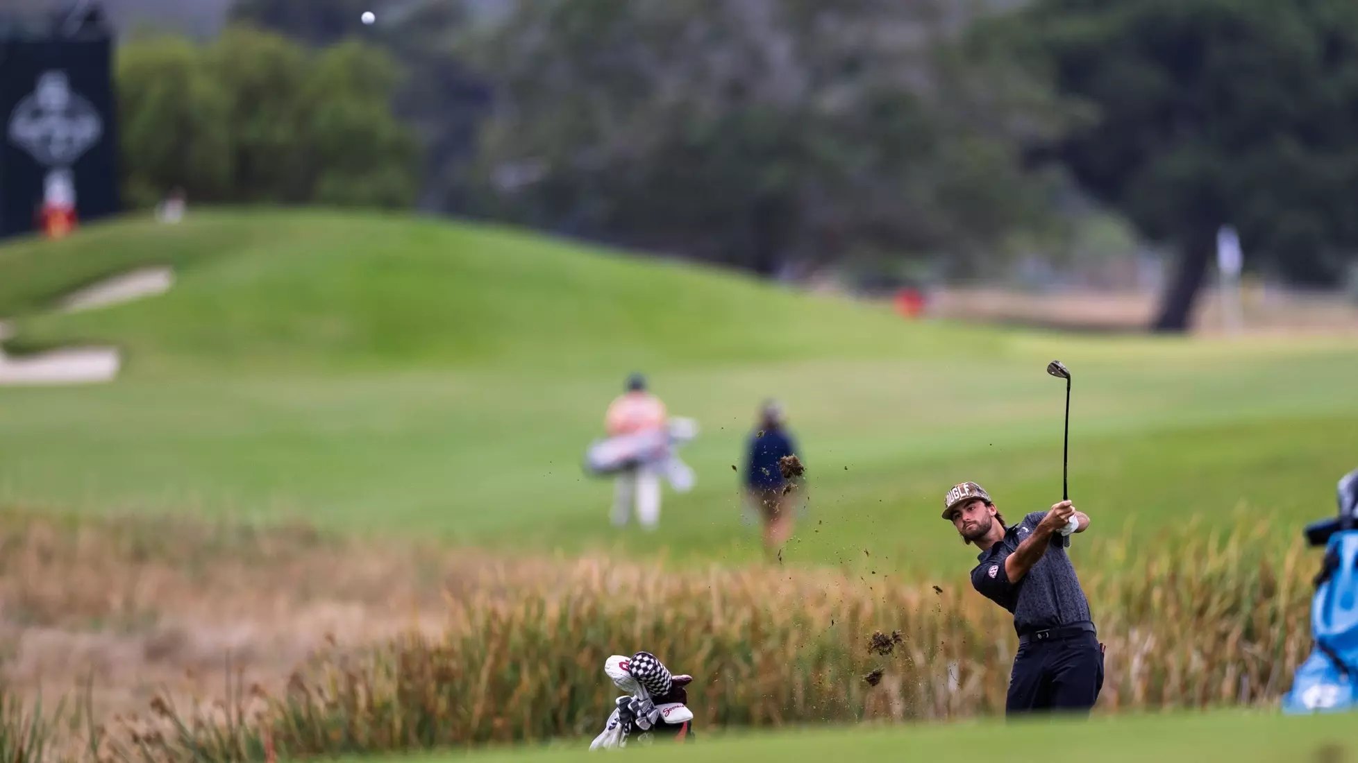 Golfer in dark clothing hits an iron shot from tall rough grass.