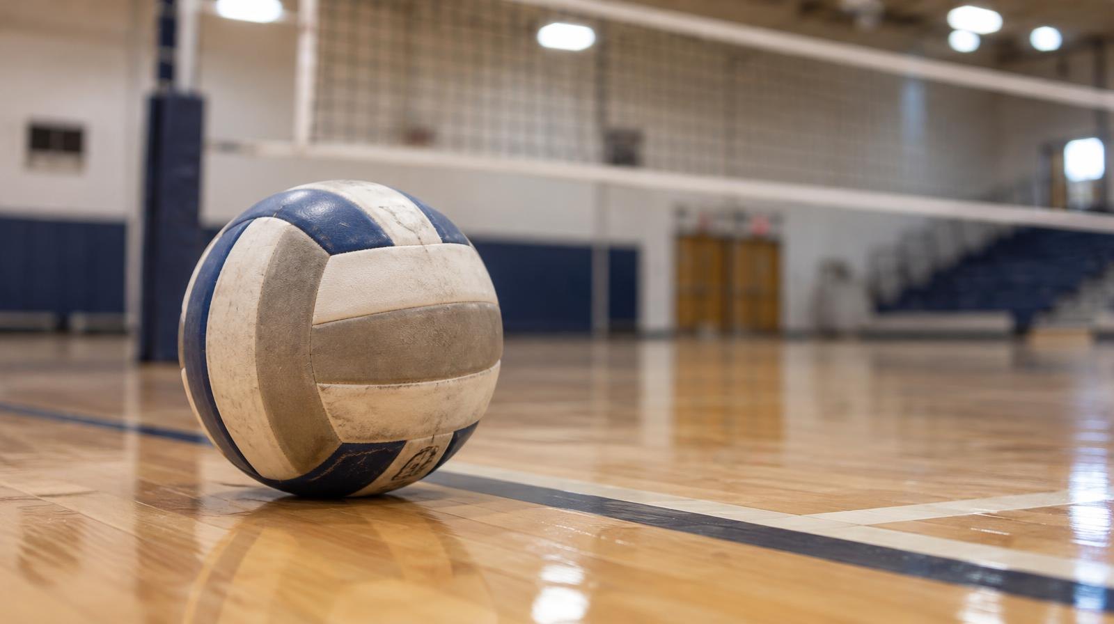 A volleyball resting on a hardwood gym floor near the net under bright overhead lights.