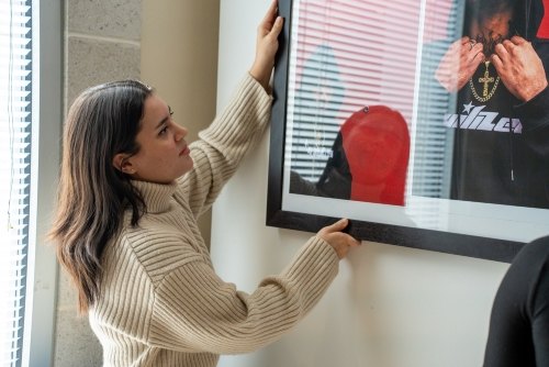 Young woman in a beige turtleneck carefully positions a framed photograph on a wall.