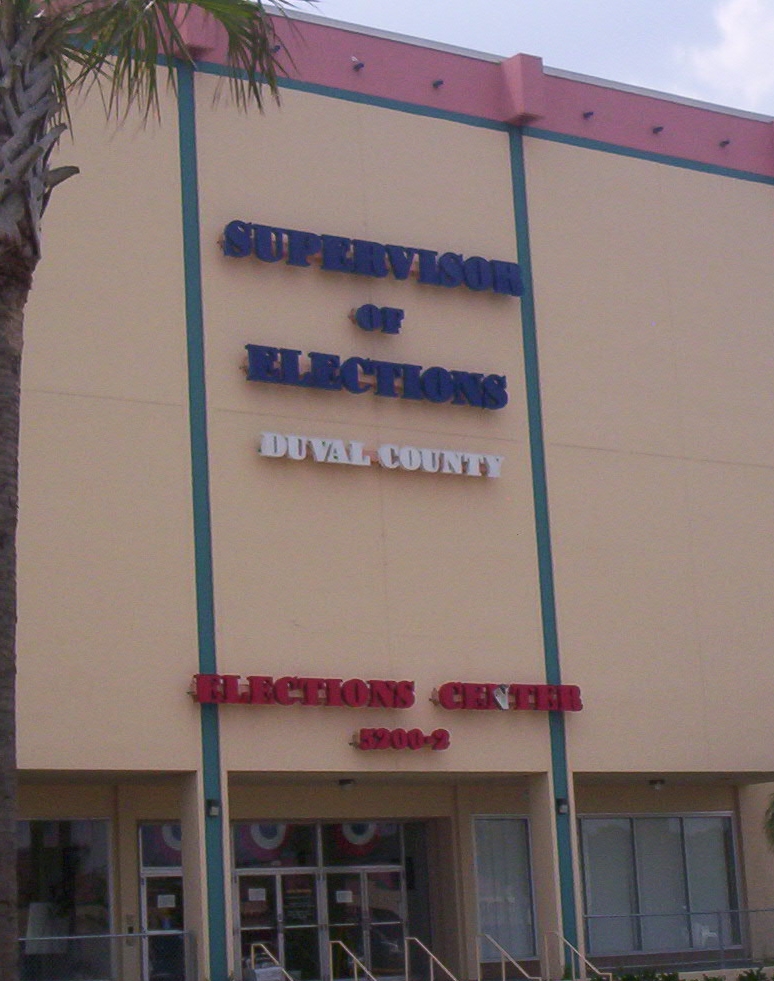 Exterior of a building with blue and red Supervisor of Elections signage and a palm tree.