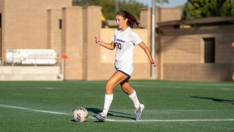 A soccer player in a white Lutheran uniform dribbles a ball on green turf.