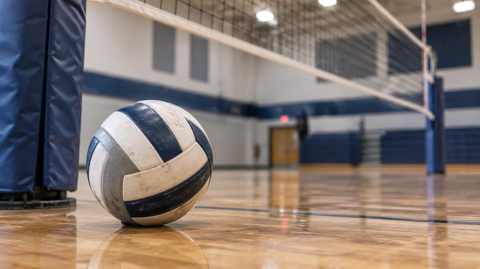 A volleyball resting on a hardwood gymnasium floor near the net post.