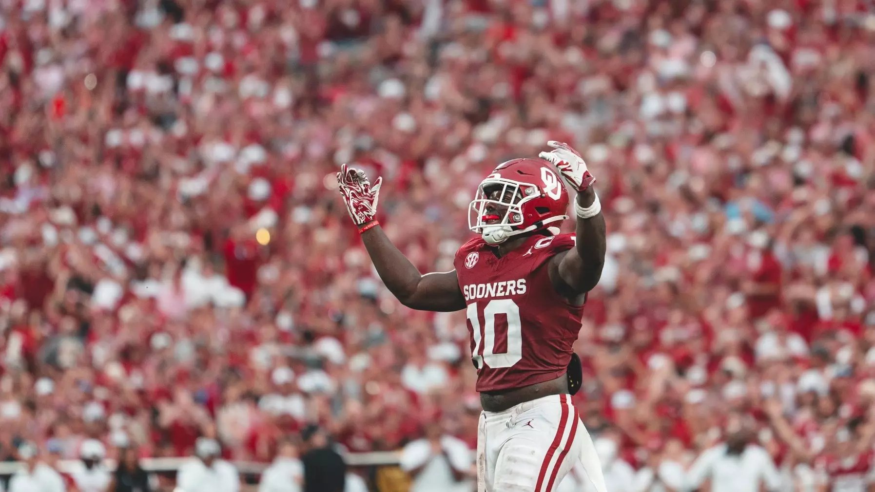 Football player in crimson jersey raises both arms in celebration before a packed stadium crowd.